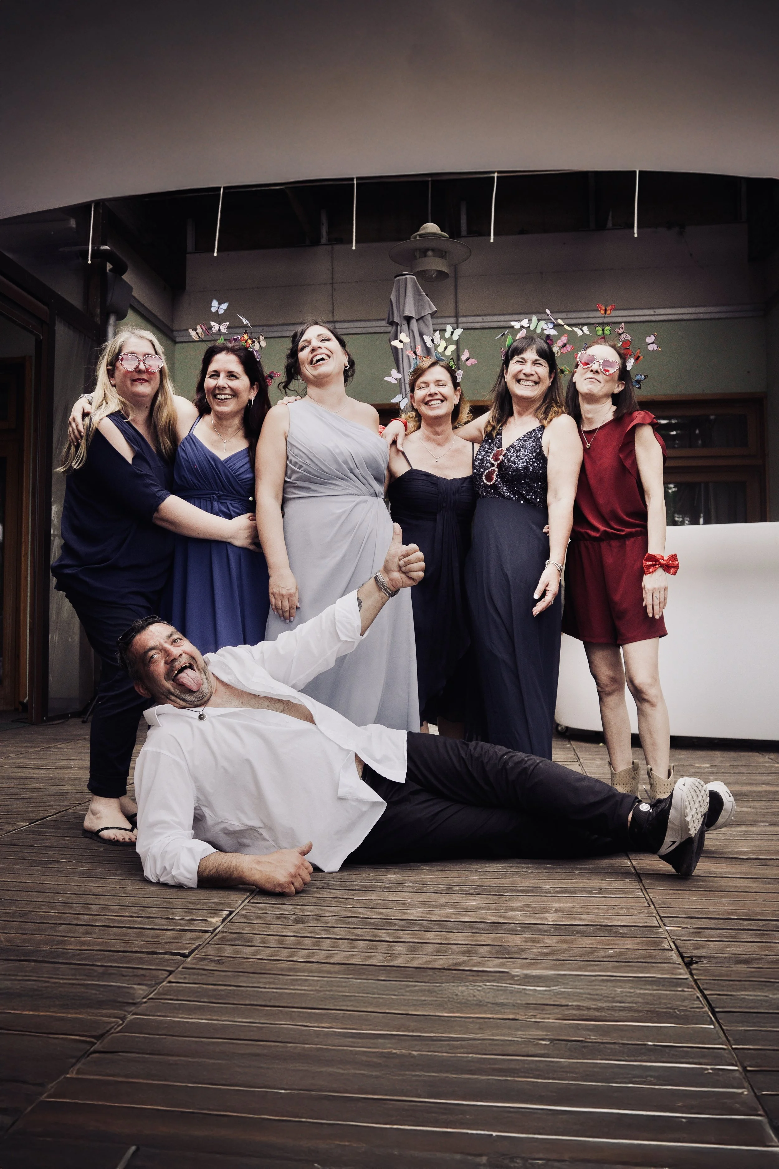 Un gruppo di sei donne e un uomo che giacciono e si trovano in piedi su un pavimento di legno, sembrano divertirsi e ridere, durante una festa o un evento sociale, con decorazioni di farfalle colorate sopra le loro teste in uno spazio interno o su una terrazza.