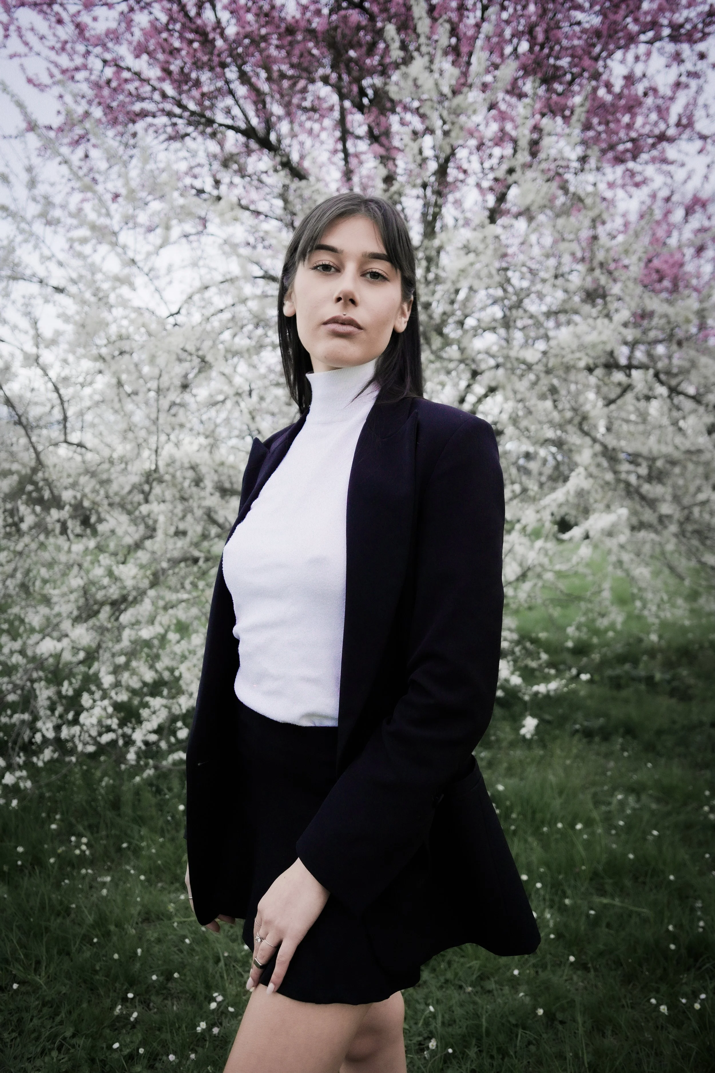 Una giovane donna in abbigliamento elegante con giacca nera e maglia bianca, che posa in un prato con alberi in fiore rosa e bianco sullo sfondo.