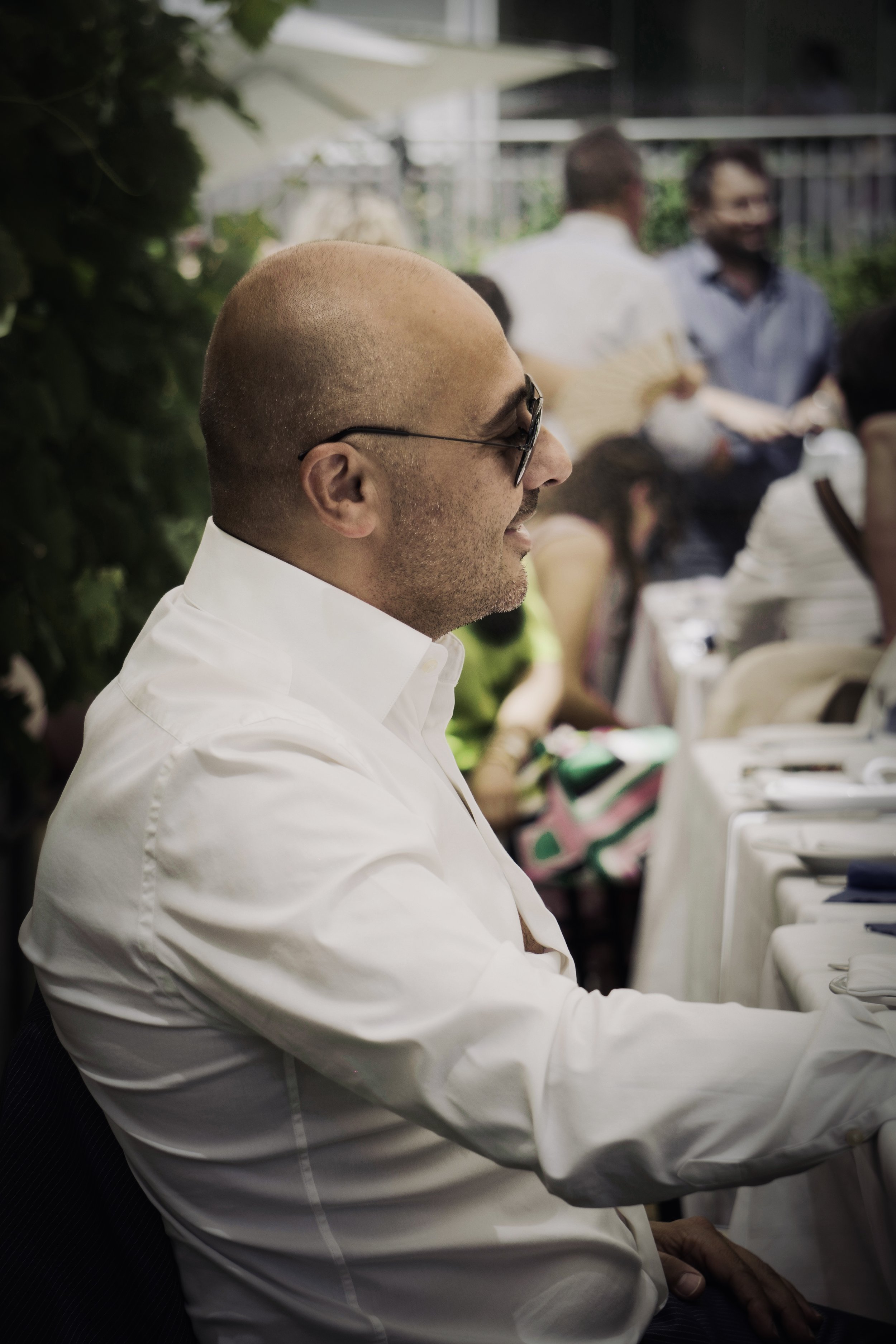 Uomo con testa rasata e occhiali da sole seduto a un tavolo in un evento sociale all'aperto, con altre persone sullo sfondo.