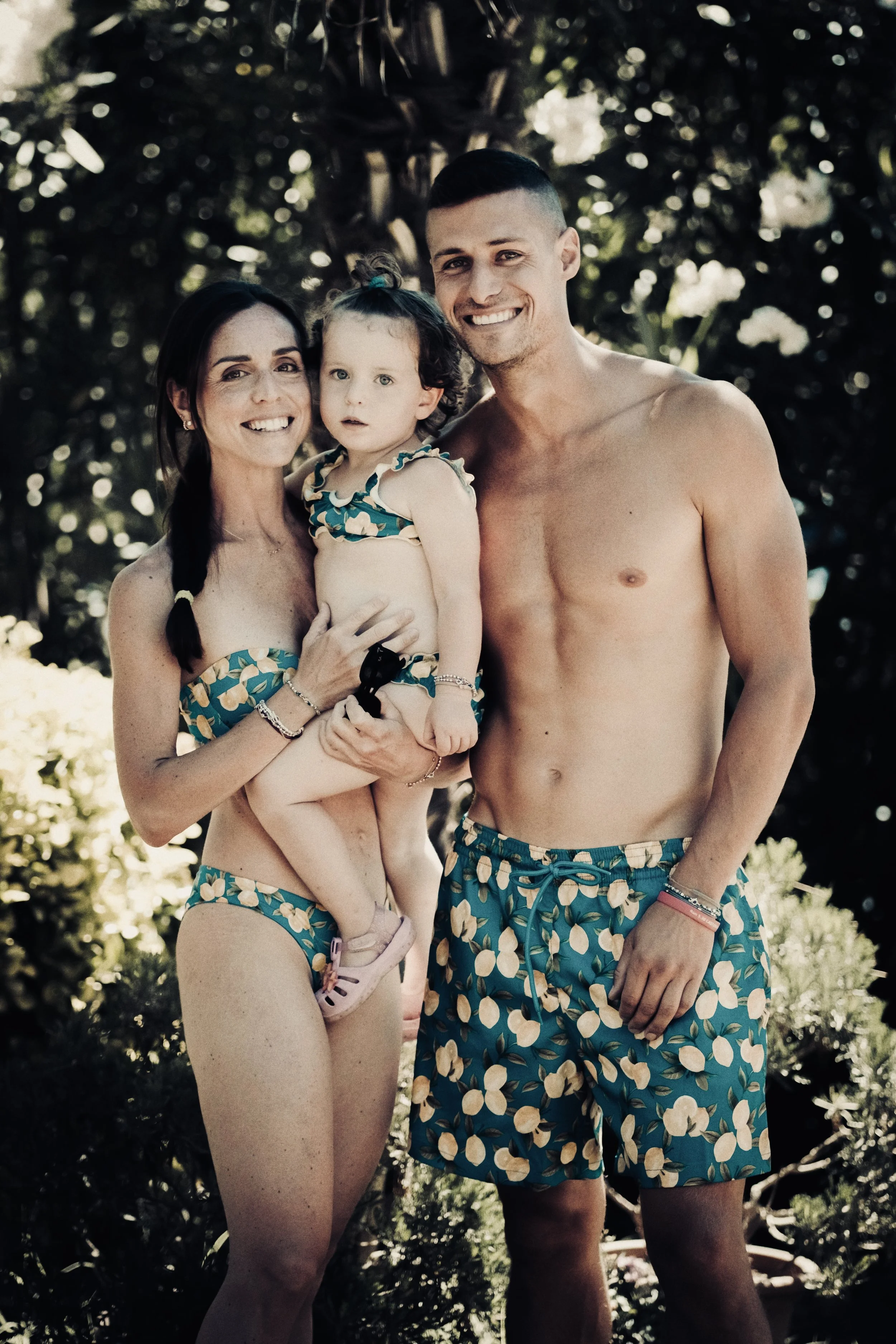 Una famiglia composta da una donna, un uomo e una bambina in abbigliamento da spiaggia con stampe di limoni, sorridenti, posano all'aperto davanti a un albero.