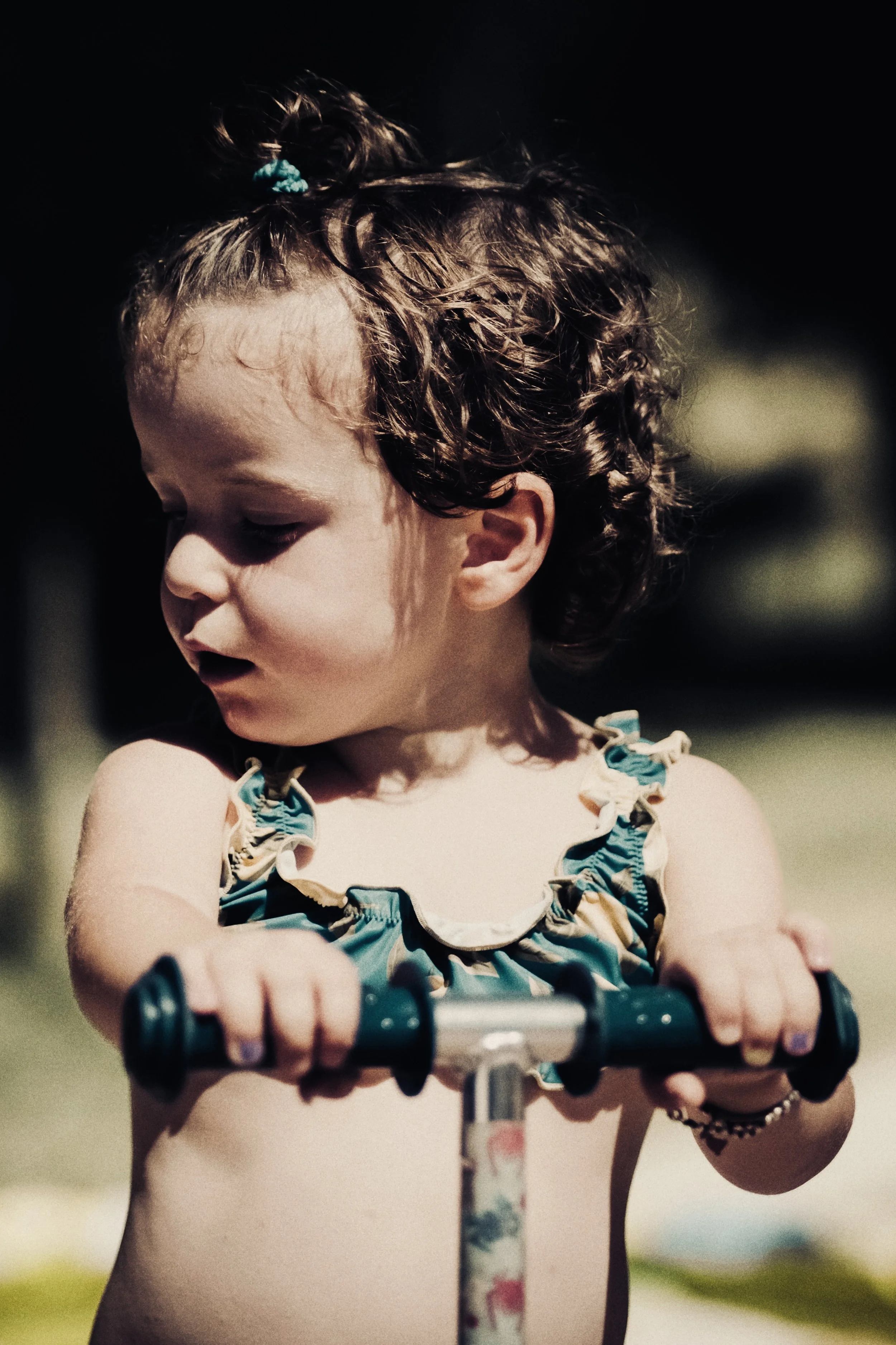 Una bambina con capelli riccioli scuri e con un fiocco blu sulla testa, che si tiene con le mani su un manubrio di biciclette o triciclo, indossa un top con spalline a volant.