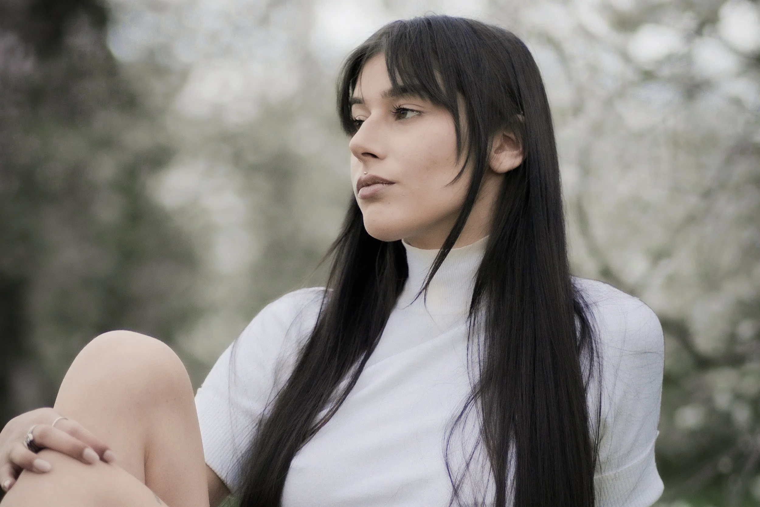 Ragazza con capelli lunghi neri, vestita di bianco, che guarda pensierosa in un paesaggio con alberi in fiore.