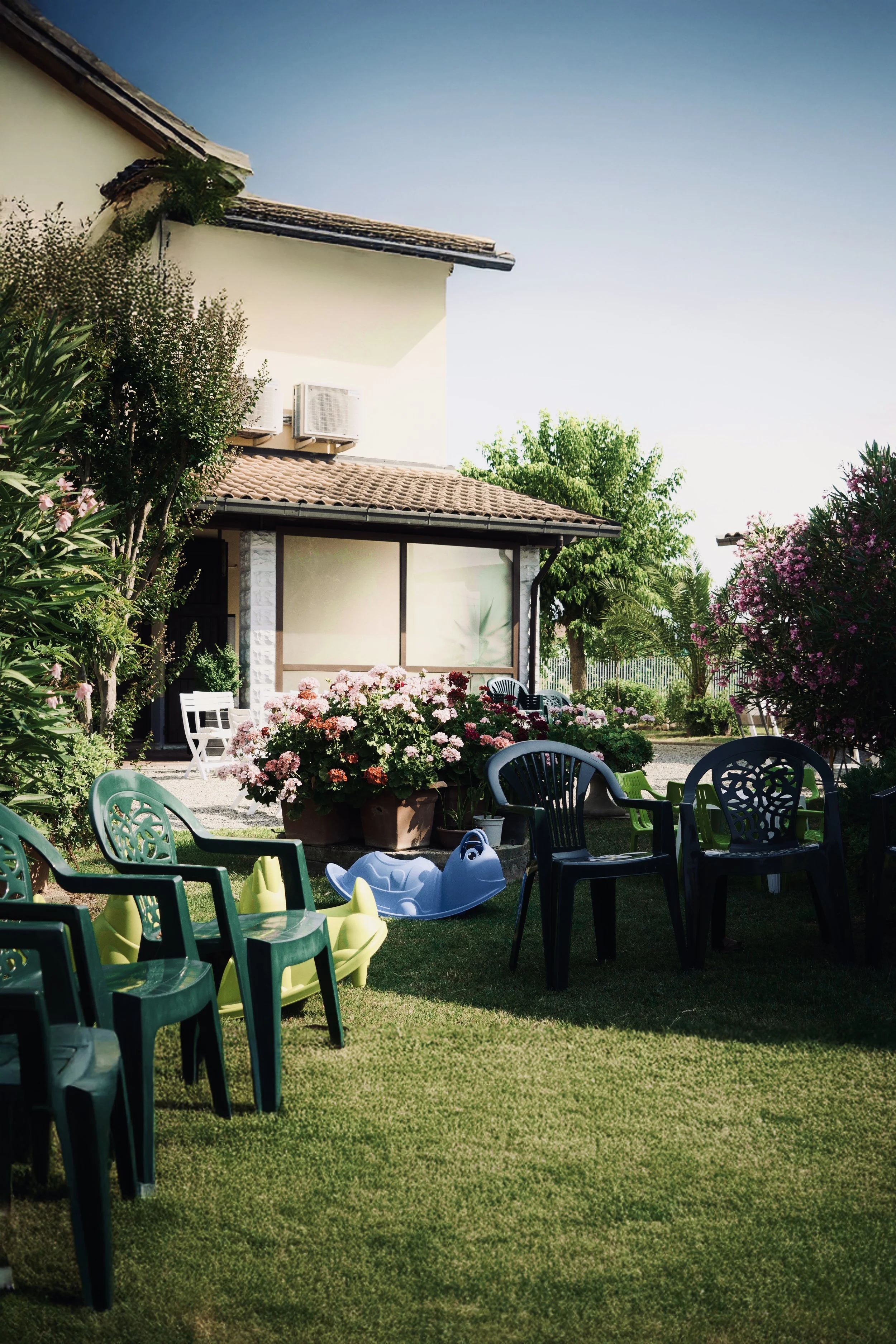 Giardino con panchine di plastica, fiori colorati e giochi per bambini, casa con aria condizionata, alberi e cielo sereno.