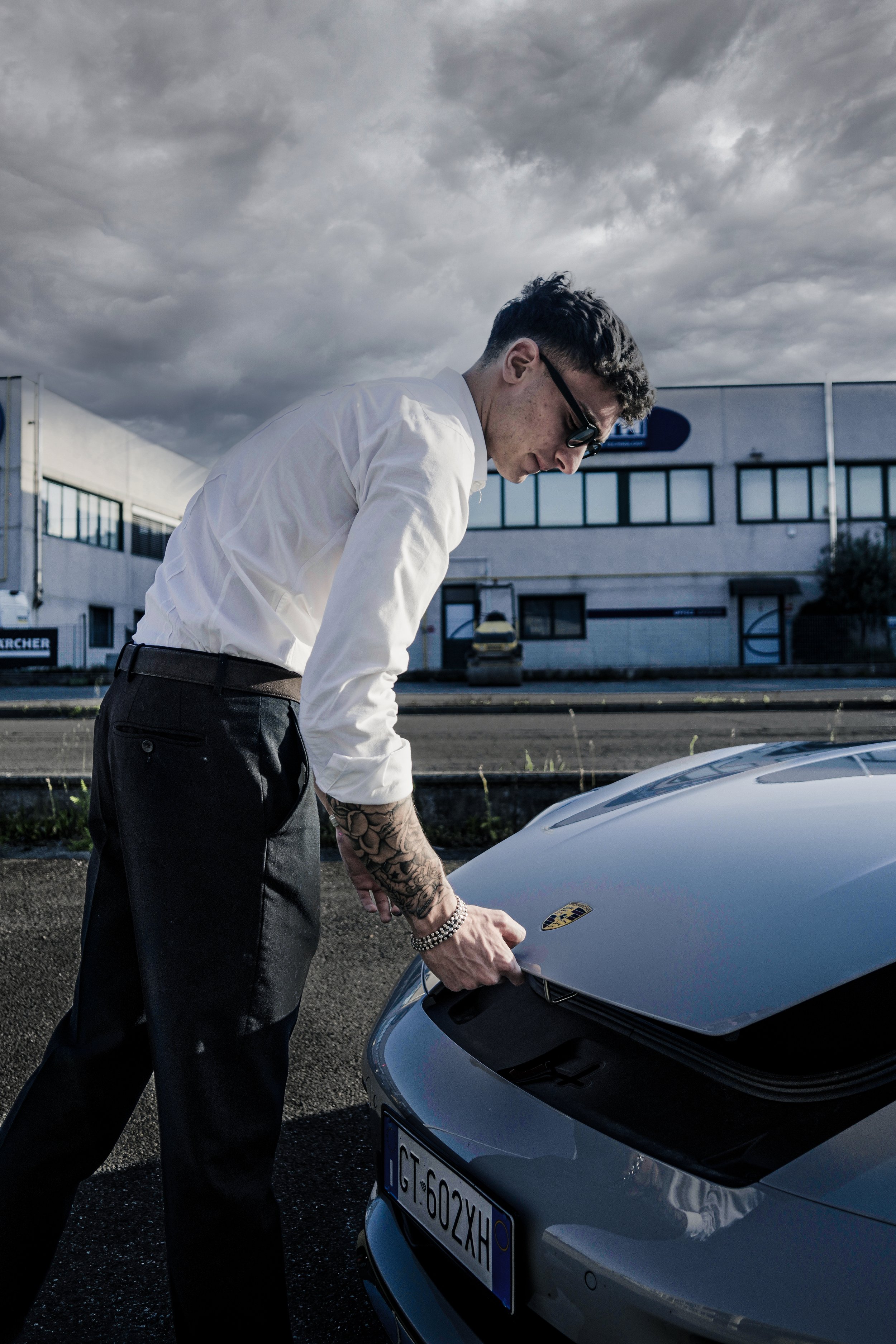 Uomo con tatuaggi, occhiali da sole, in camicia bianca e pantaloni neri, vicino a una Porsche grigia in un parcheggio