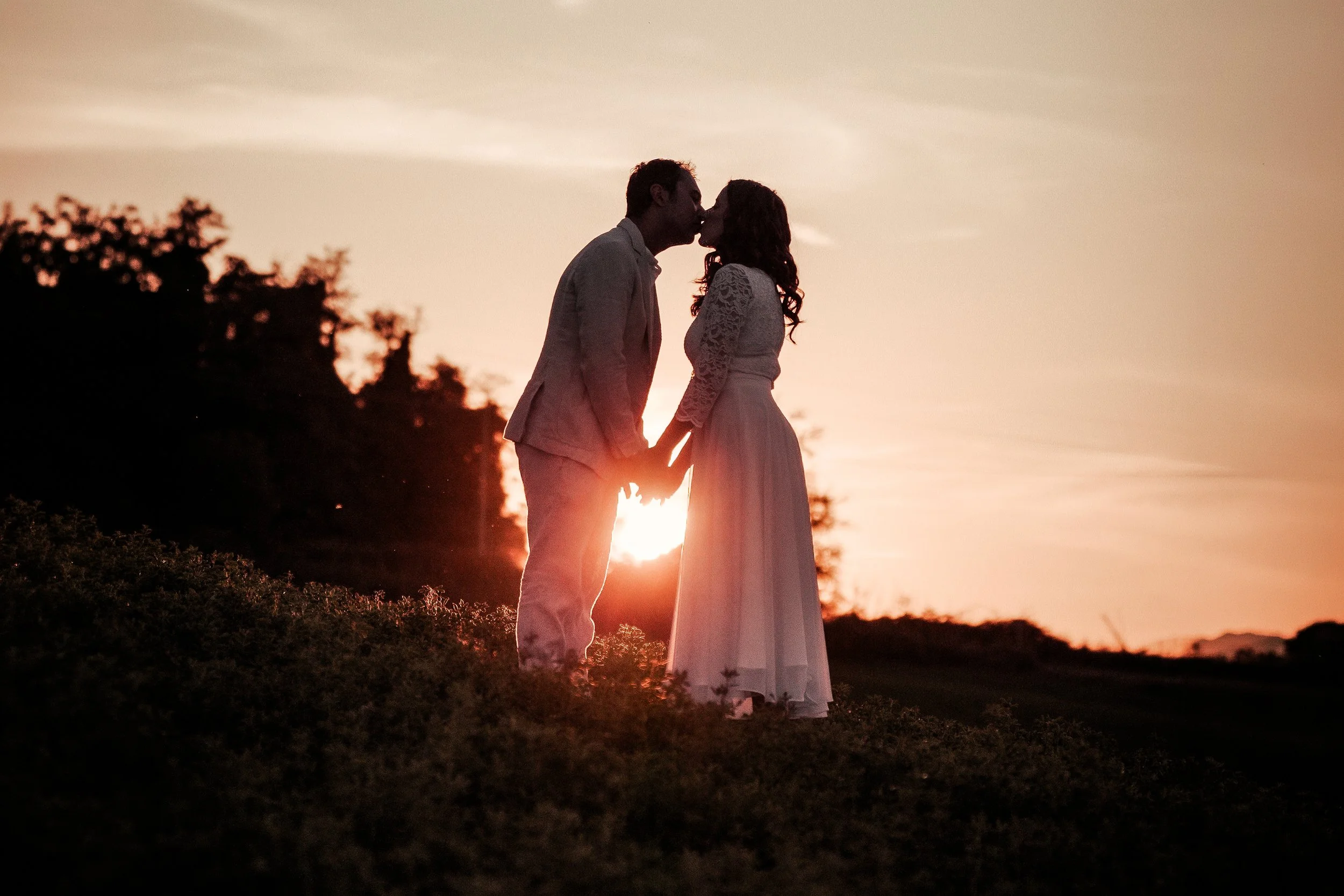 Coppia di sposi che si bacia al tramonto, con sfondo un cielo arancione e silhouette di alberi.