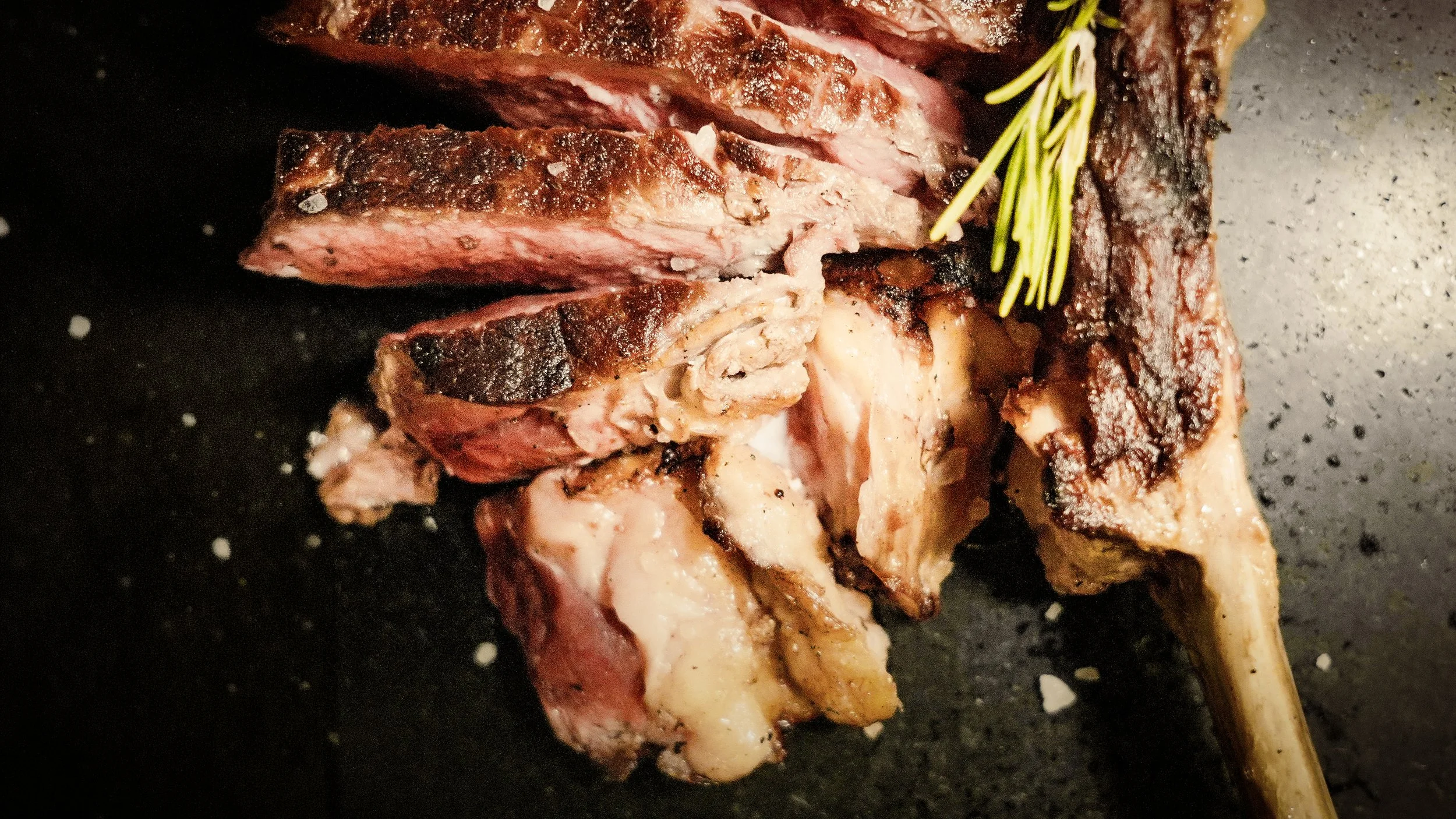 Una bistecca alla griglia con il midollo visibile, decorata con un rametto di rosmarino, su una superficie nera.