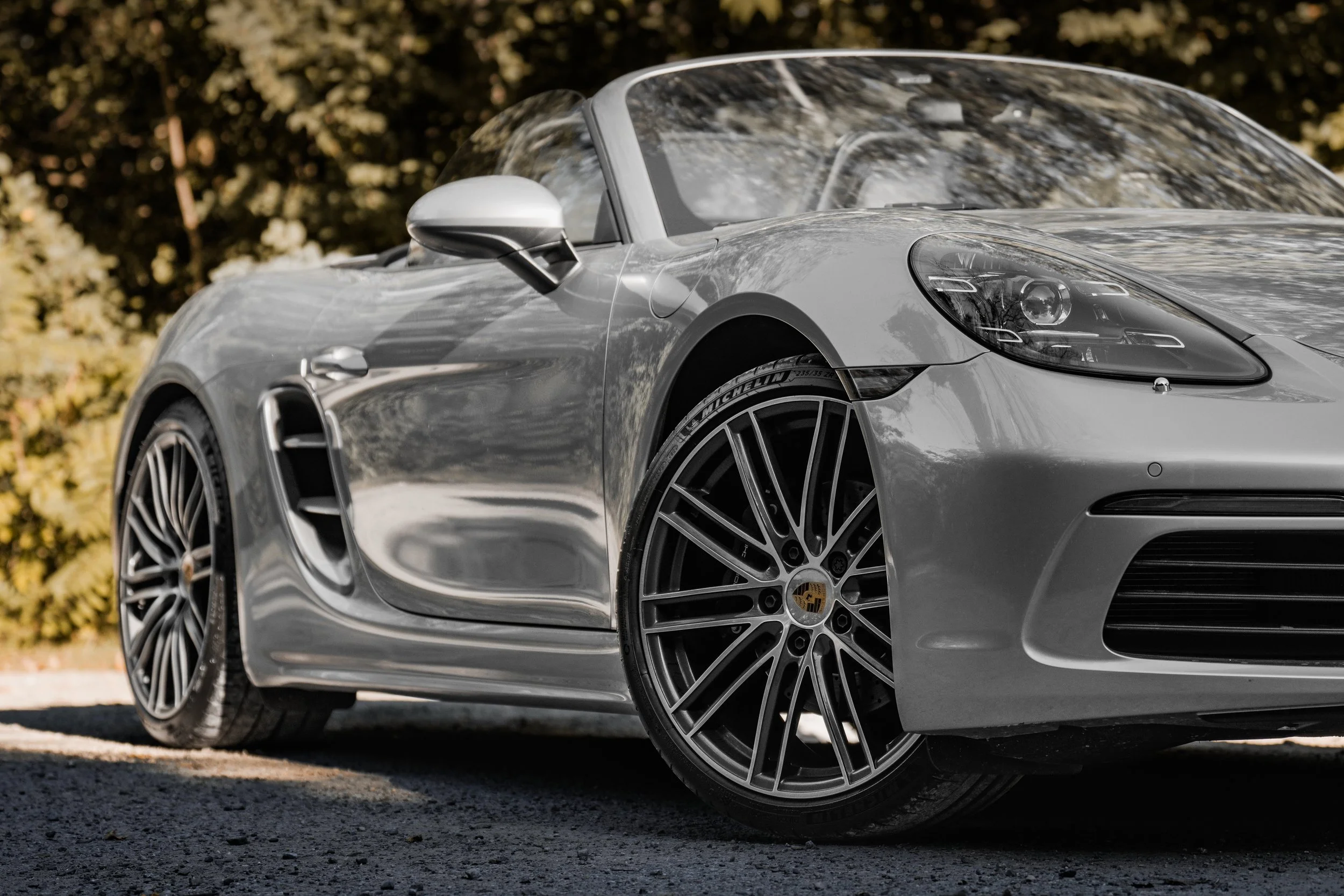 Auto sportiva Porsche gris, vista frontale e laterale, con cerchi in lega e luci accese, parcheggiata su strada con alberi sullo sfondo.