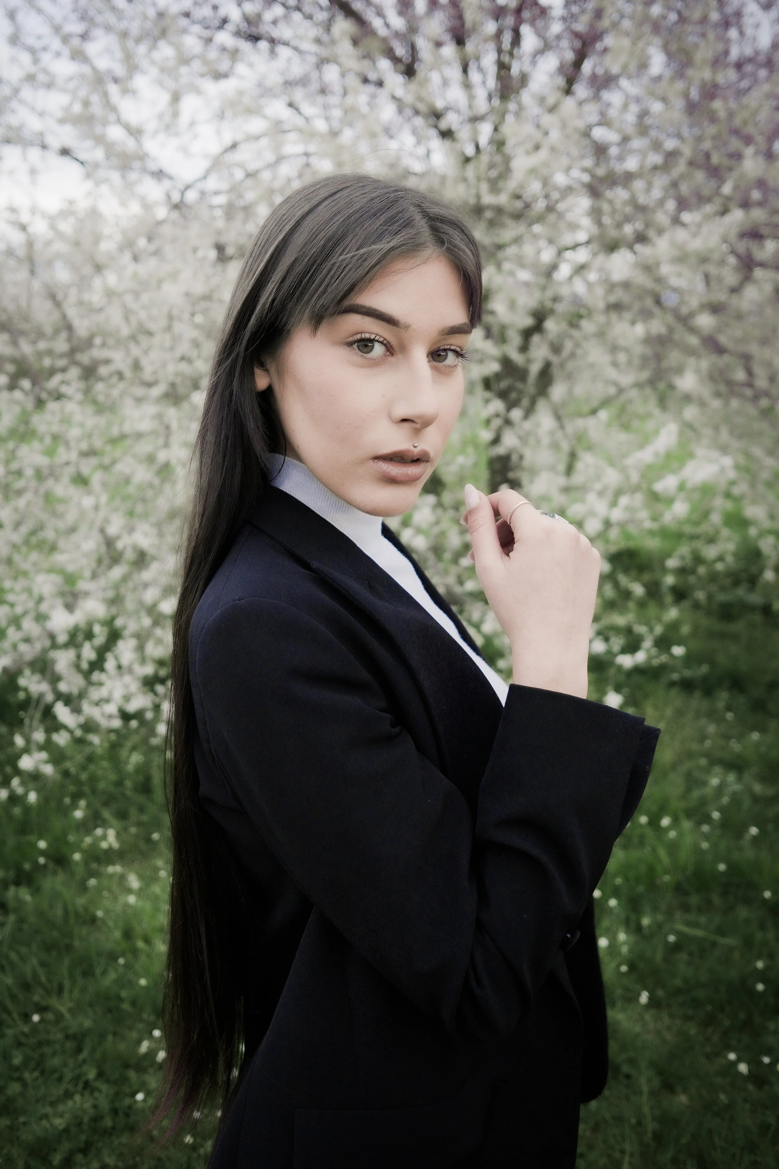 Ragazza con capelli lunghi e occhi chiari, vestita con un blazer nero e una camicia bianca, in piedi in un giardino con alberi in fiore, vestita elegantemente.