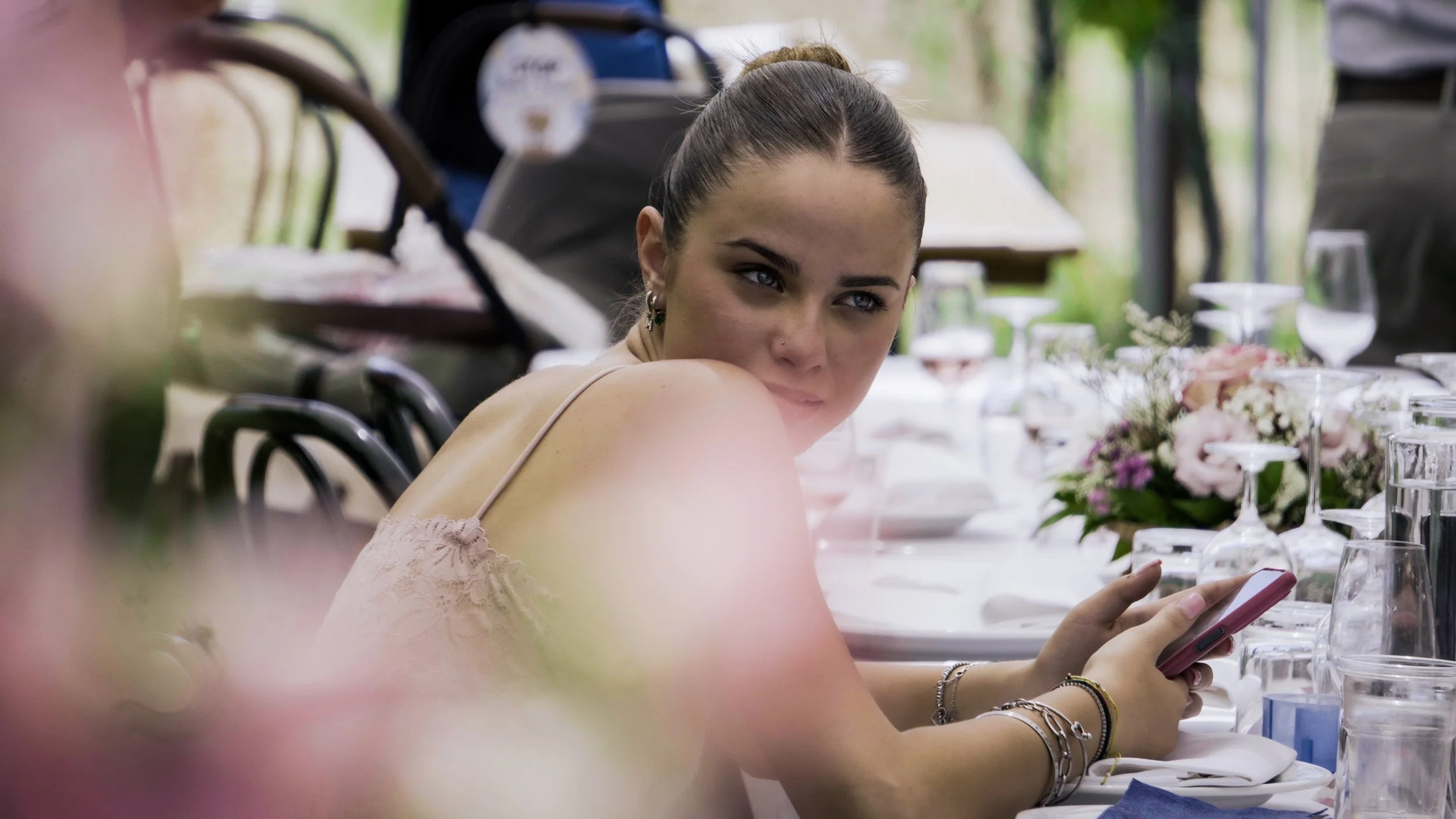 Una giovane donna con capelli raccolti, indossa una cima leggera di colore beige, seduta a un tavolo apparecchiato con bicchieri, piatti, posate e un centrotavola con fiori rosa e bianchi, mentre guarda il telefono cellulare.