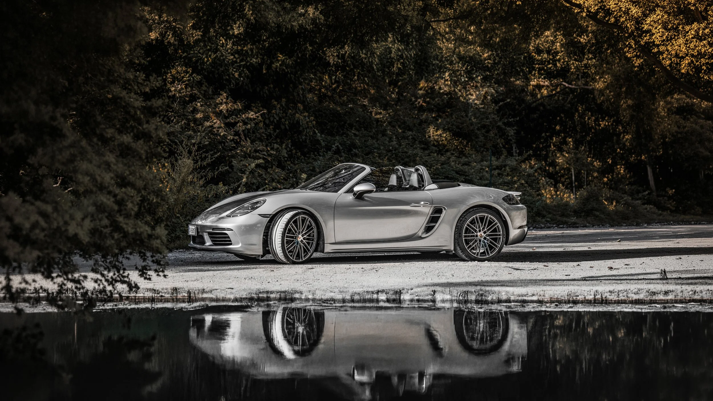 Una macchina sportiva convertibile di colore grigio è parcheggiata su una strada circondata da alberi con foglie verdi e gialle, con un riflesso della macchina nell'acqua sulla strada.