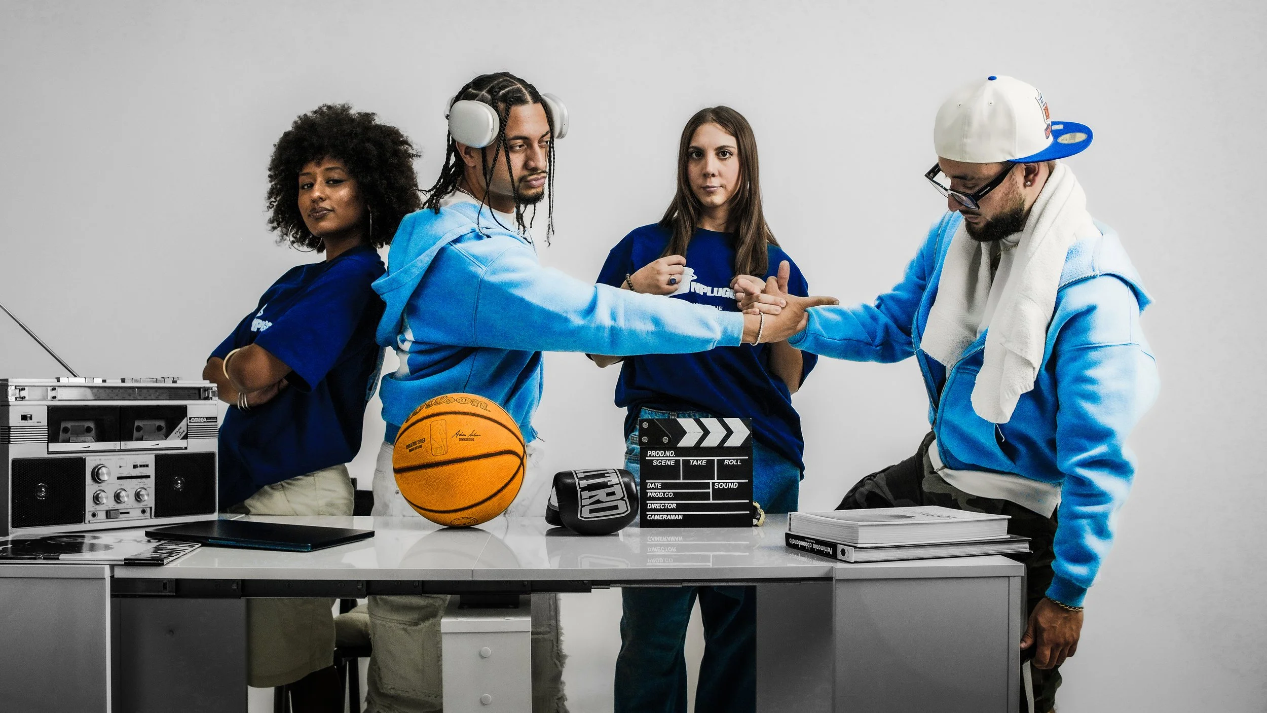 Quattro giovani uomini e donne di diverse etnie in piedi dietro un tavolo bianco in uno studio con sfondo bianco, con attrezzature cinematografiche e sportivi come un pallone da basket, una cuffia, un'imbottitura e una cartella con una sceneggiatura, mentre si scambiano un'alta mano