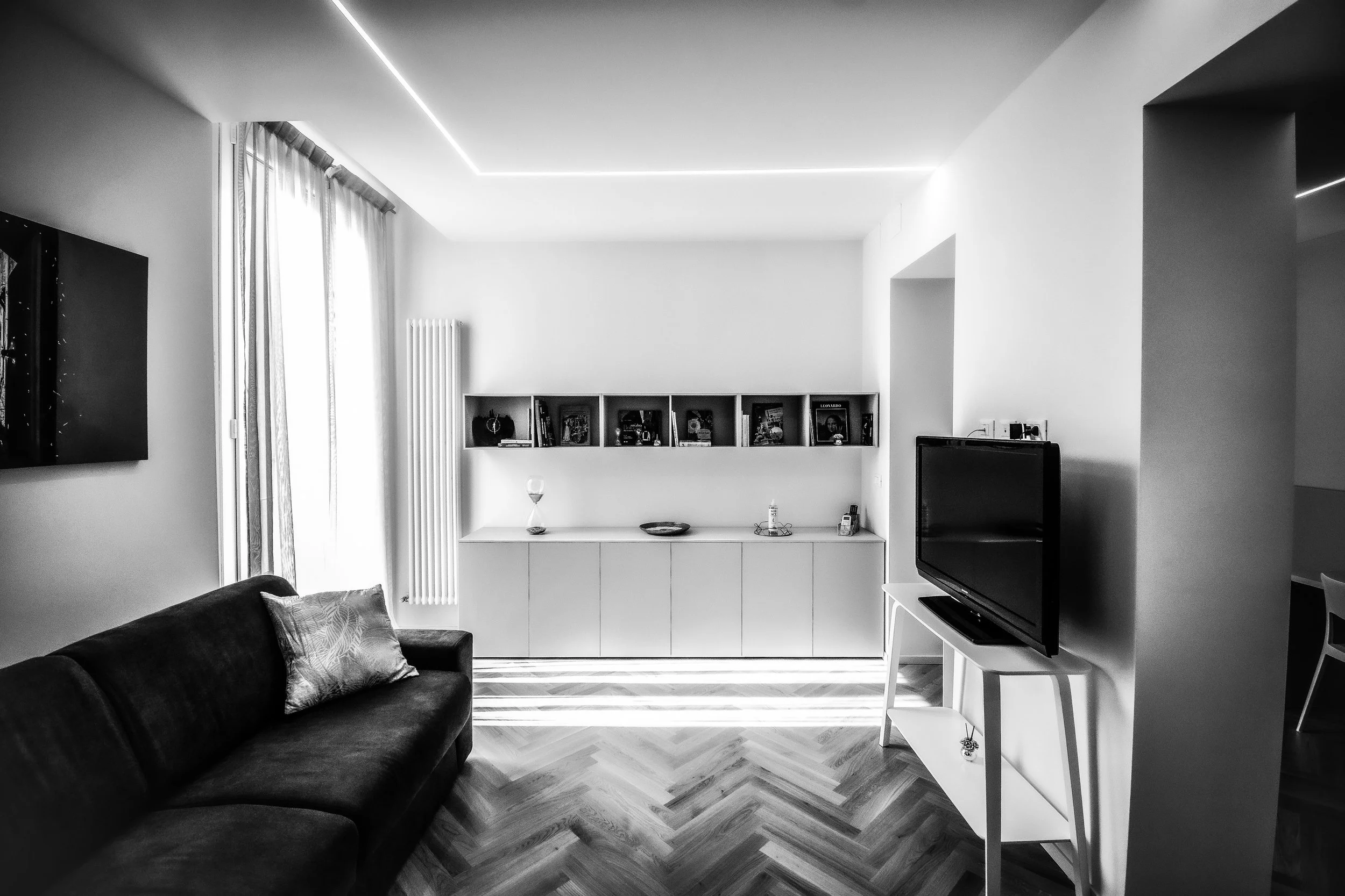 Interno di un soggiorno moderno con divano nero, televisore su mobile bianco, libreria con libri e decorazioni, pareti bianche, pavimento a spina di pesce, tendaggi leggeri e luci a led nel soffitto.