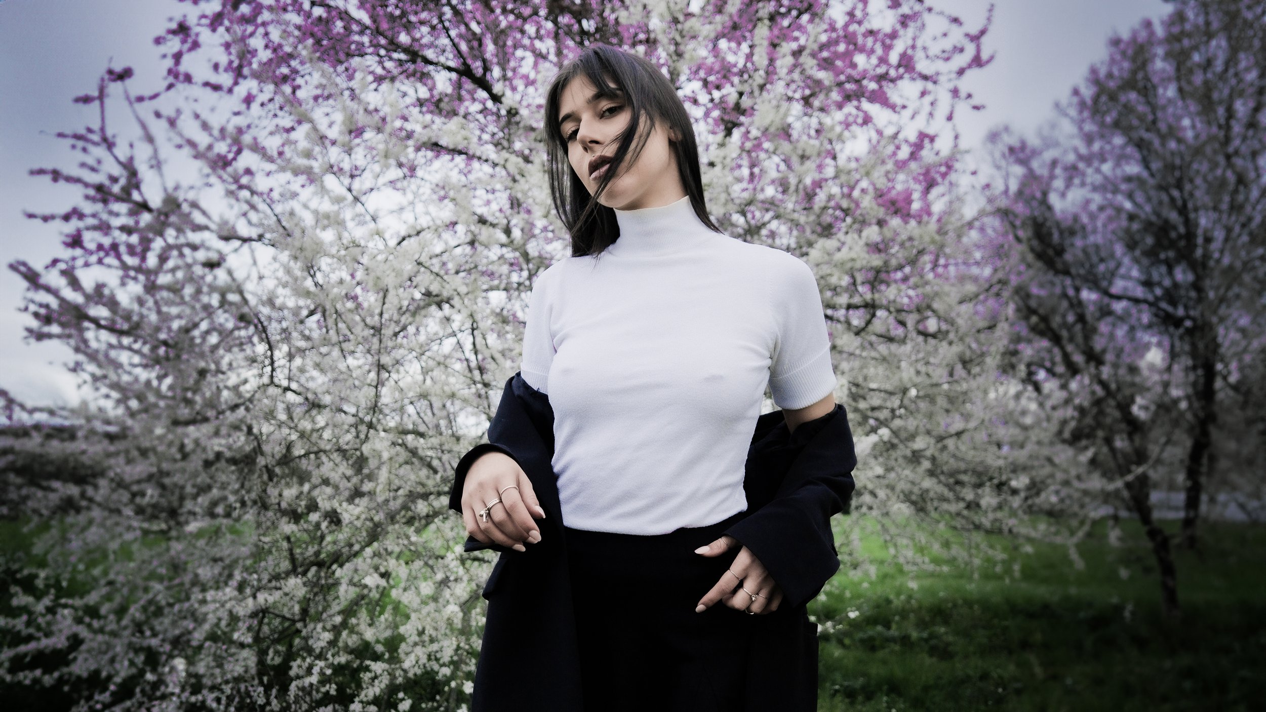 Una donna con capelli neri e trucco scuro si trova davanti a un albero in fiore con fiori rosa e bianchi. Indossa una maglietta bianca a maniche corte, una giacca scura e una gonna nera.
