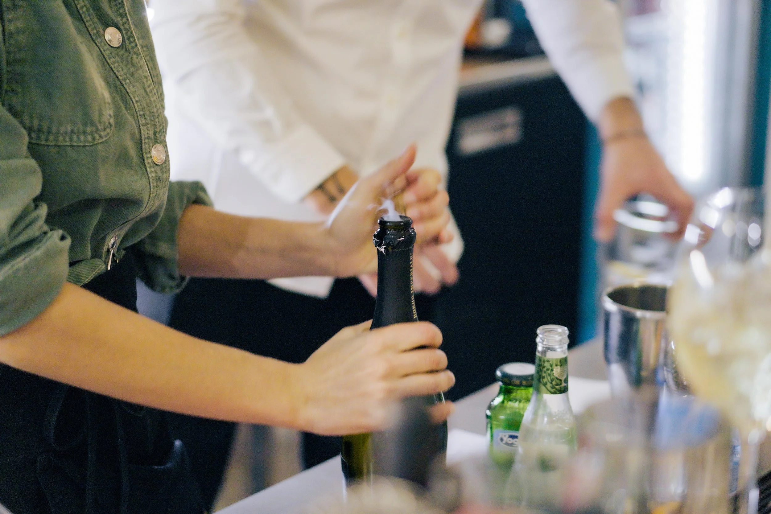 Persone che aprono una bottiglia di champagne con il tappo che scoppia, in un ambiente di festa con altre bevande e bicchieri sul tavolo.