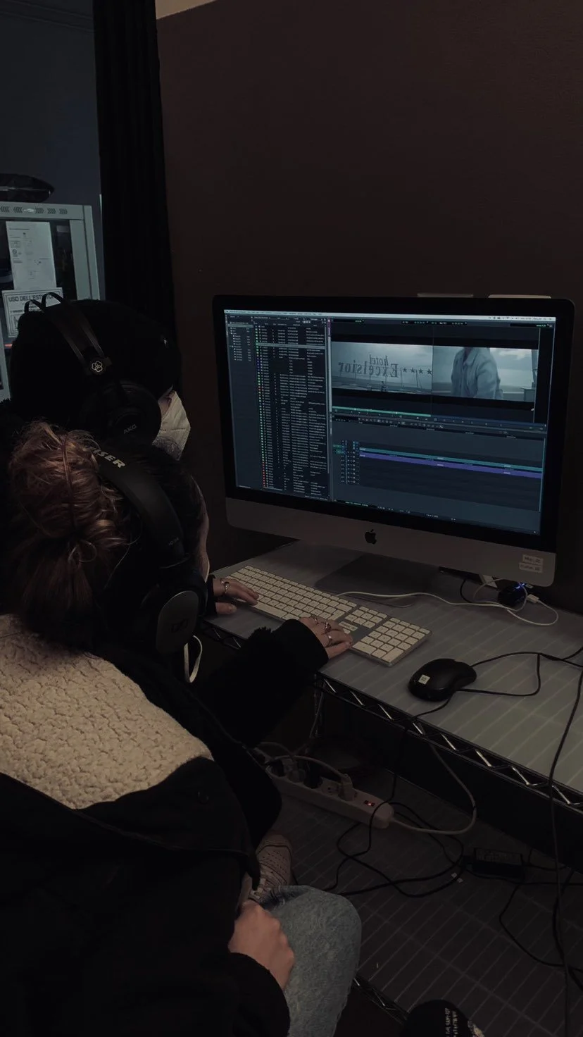 Immagine di due persone che lavorano a un computer con un software di editing video nella scena. Una persona indossa cuffie e sta digitando sulla tastiera, mentre l'altra, seduta accanto, è visibile solo parzialmente con scarpe sportive. Sul desktop si vede un monitor con un progetto di editing video aperto.