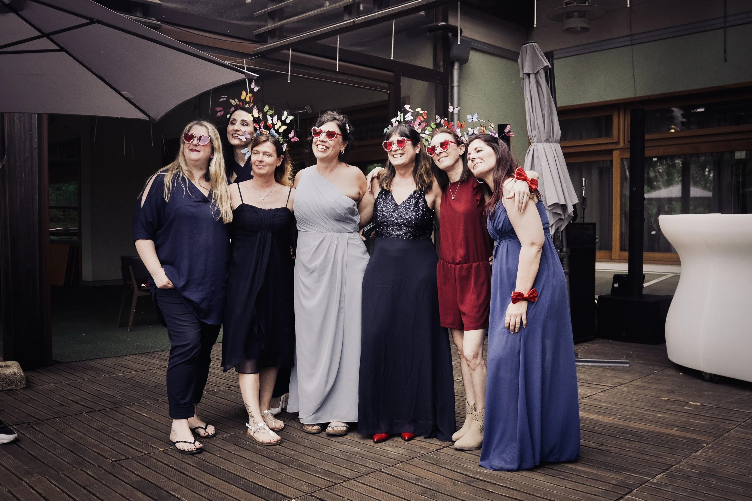 Gruppo di donne sorridenti con occhiali a cuore e decorazioni di farfalle, in posa all'aperto in un ambiente coperto.