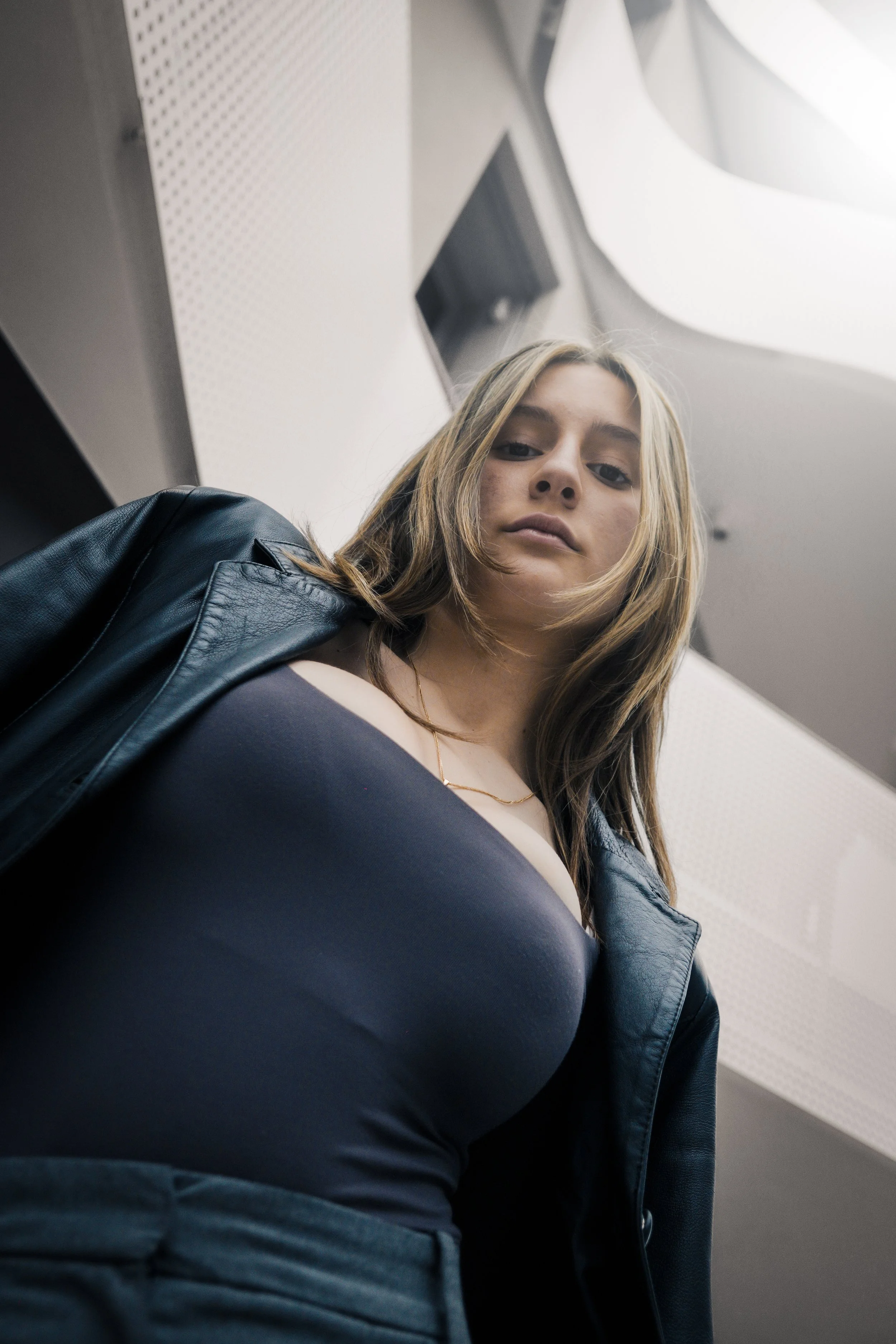 Una giovane donna vista dall'alto, che indossa una camicetta nera e una giacca di pelle, con i capelli castani chiari e lo sguardo direttamente verso la fotocamera in un ambiente con soffitto bianco e luci artificiali.