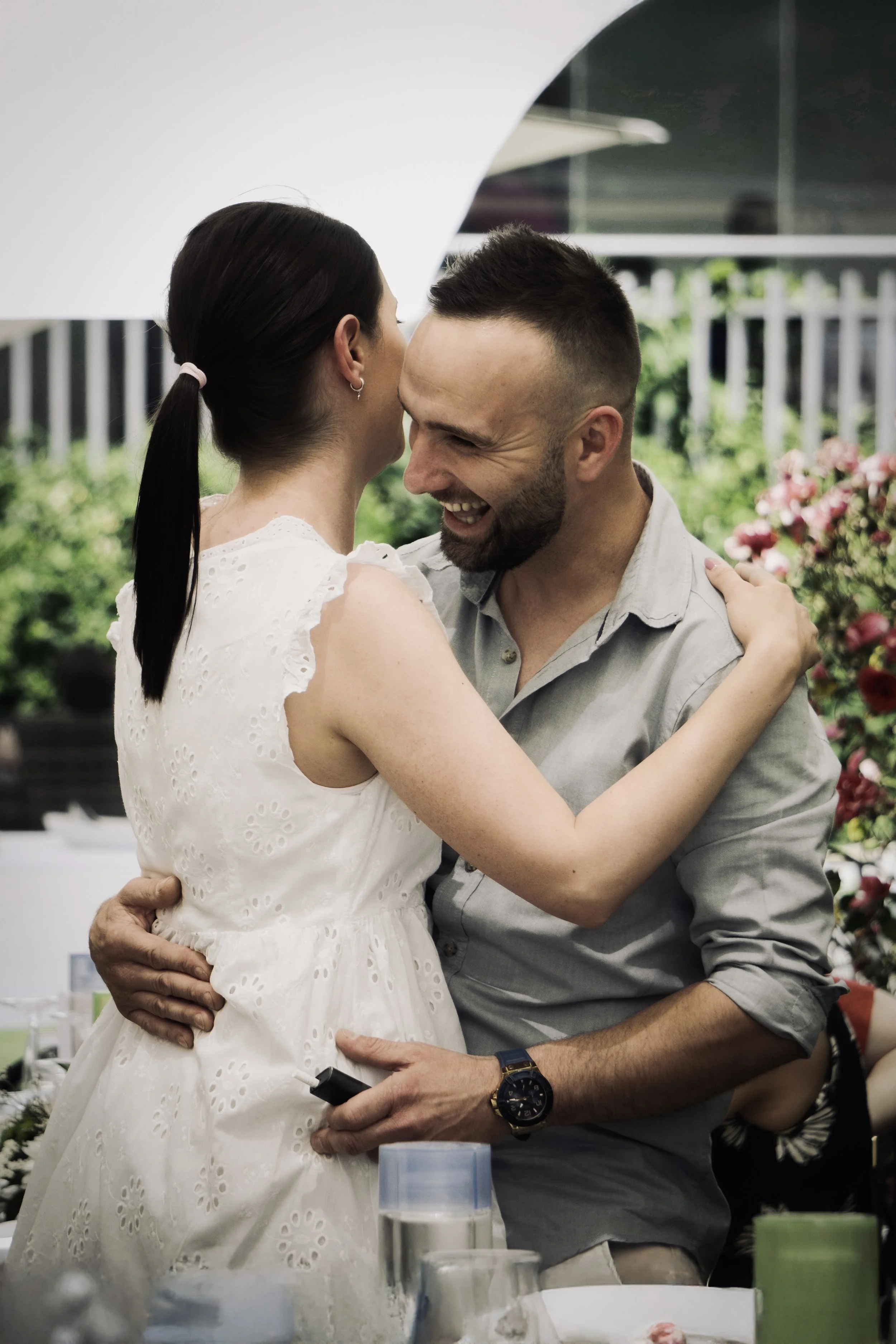 Una coppia felice si abbraccia e si bacia in un contesto di festa, presumibilmente un matrimonio, in un ambiente con piante e fiori sullo sfondo.