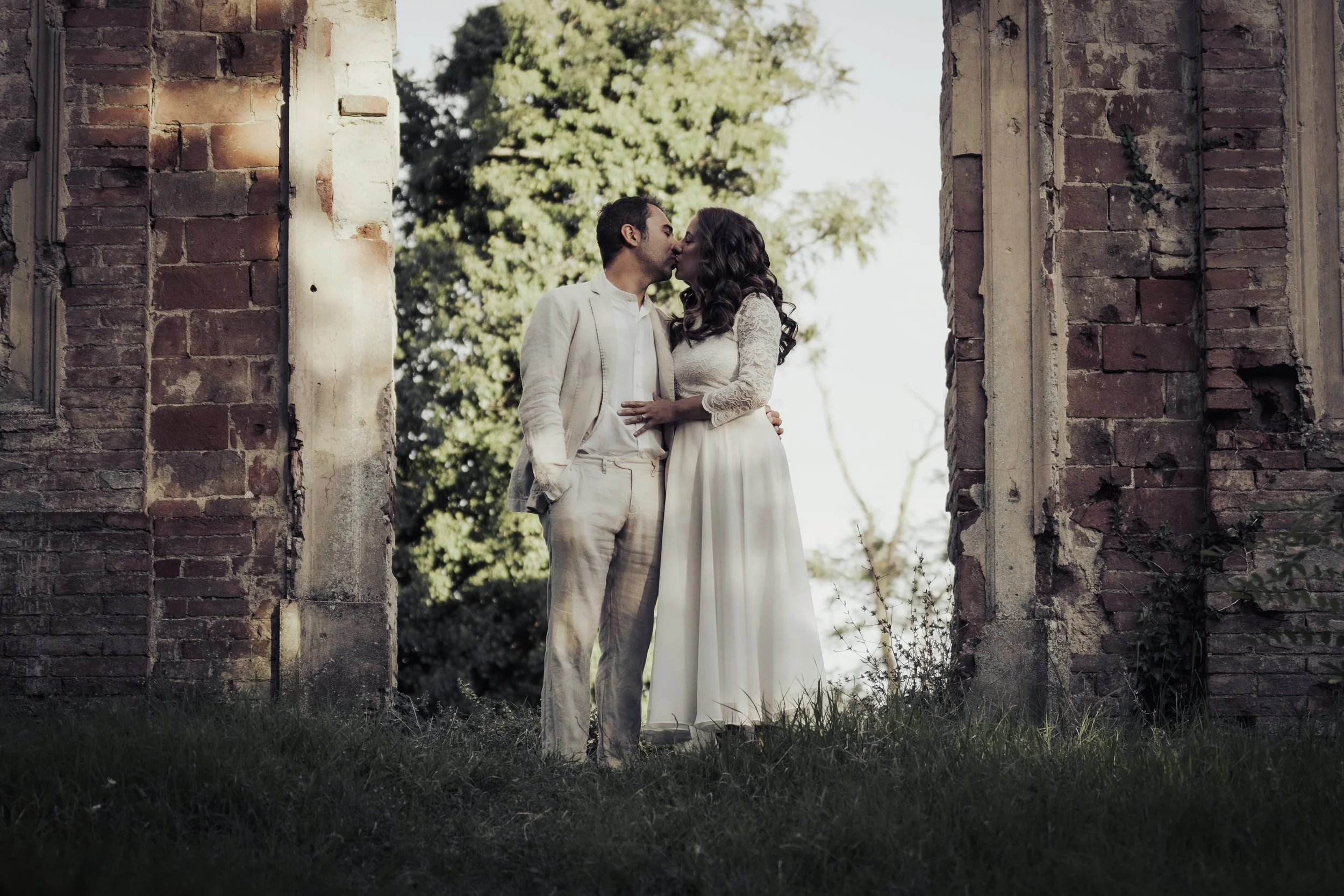 Una coppia innamorata in abiti da matrimonio si bacia sotto un arco di mattoni antichi, con sfondo di alberi e cielo.