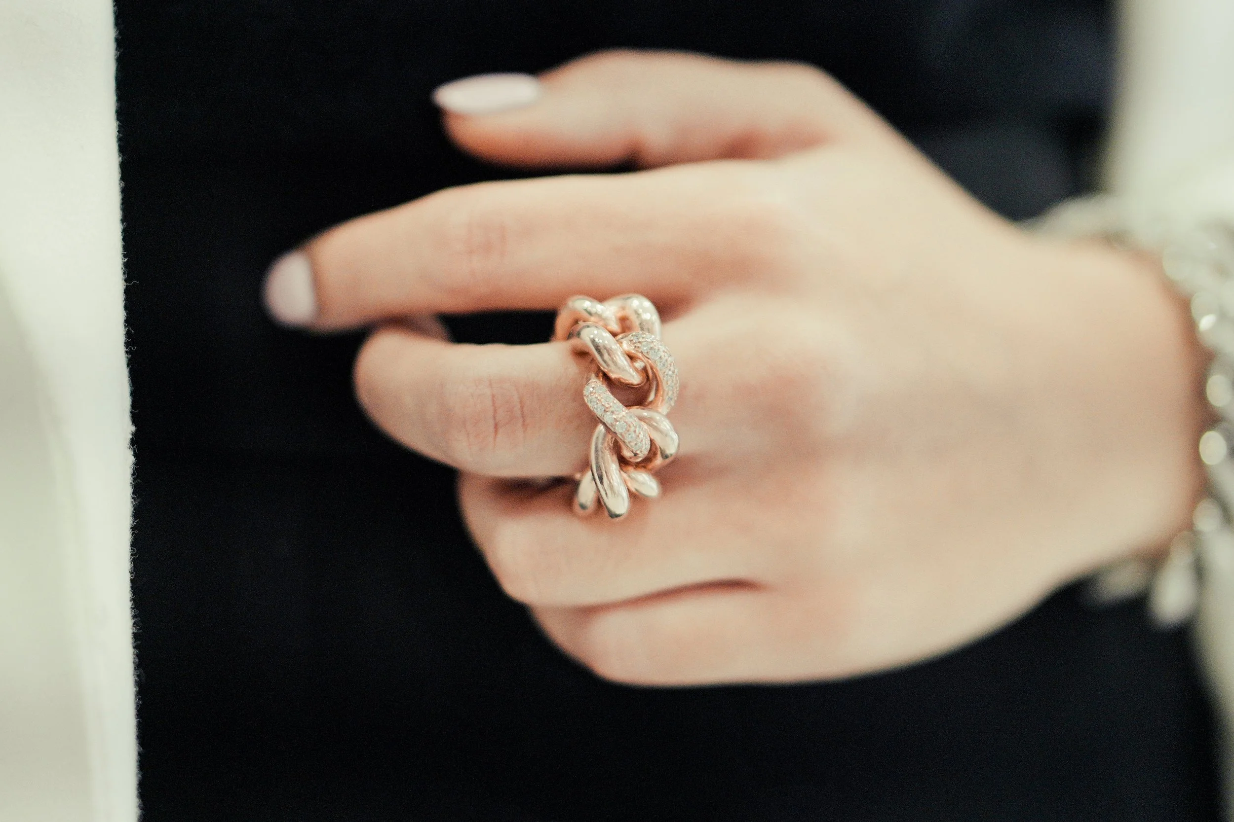 Dito di una mano indossa un anello composto da catene di metallo, alcune con dettagli di pietre preziose, con sfondo nero.