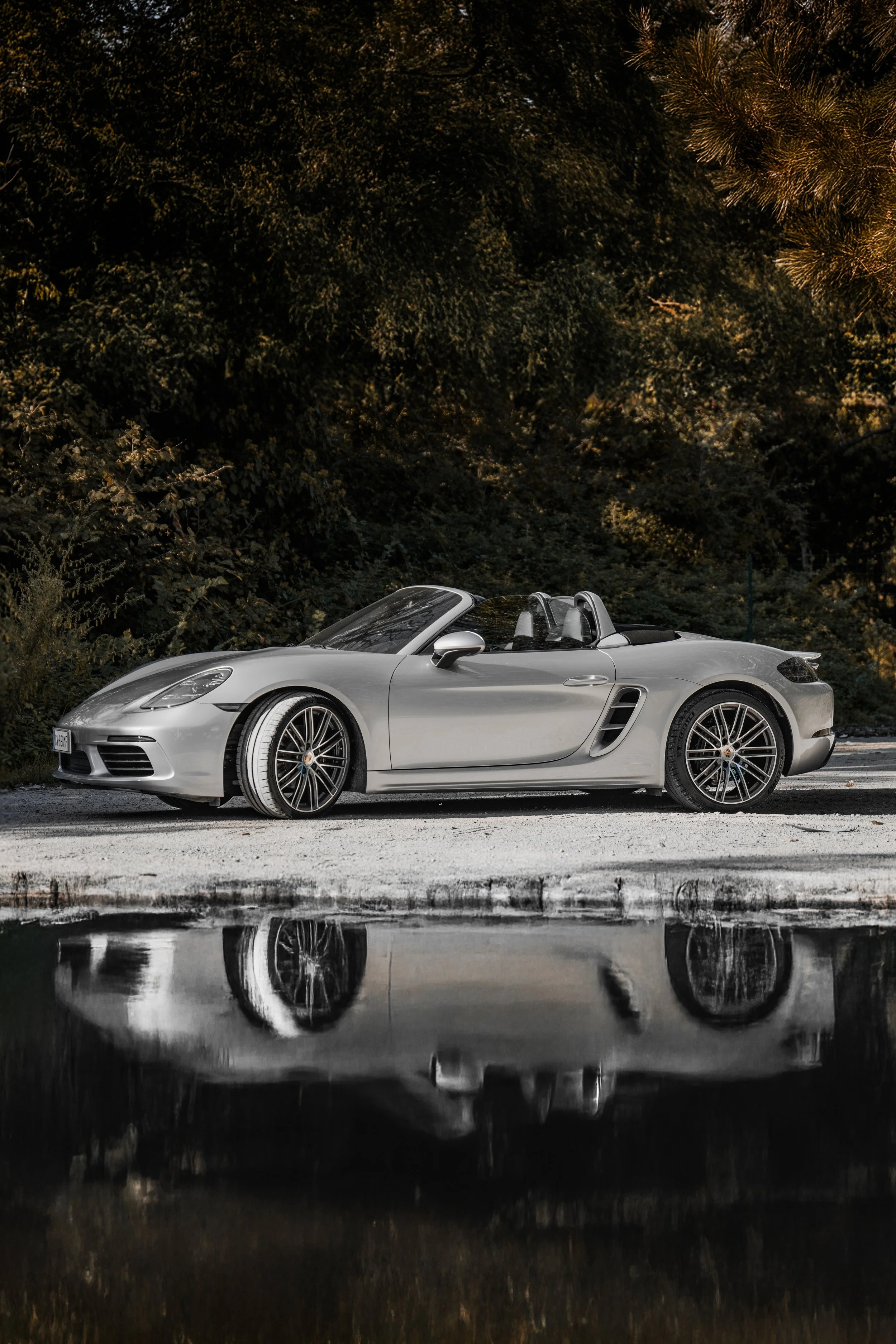 Una Mercedes-Benz AMG GT Cabrio color argento parcheggiata su una strada vicino a un laghetto, con alberi e foresta sullo sfondo.
