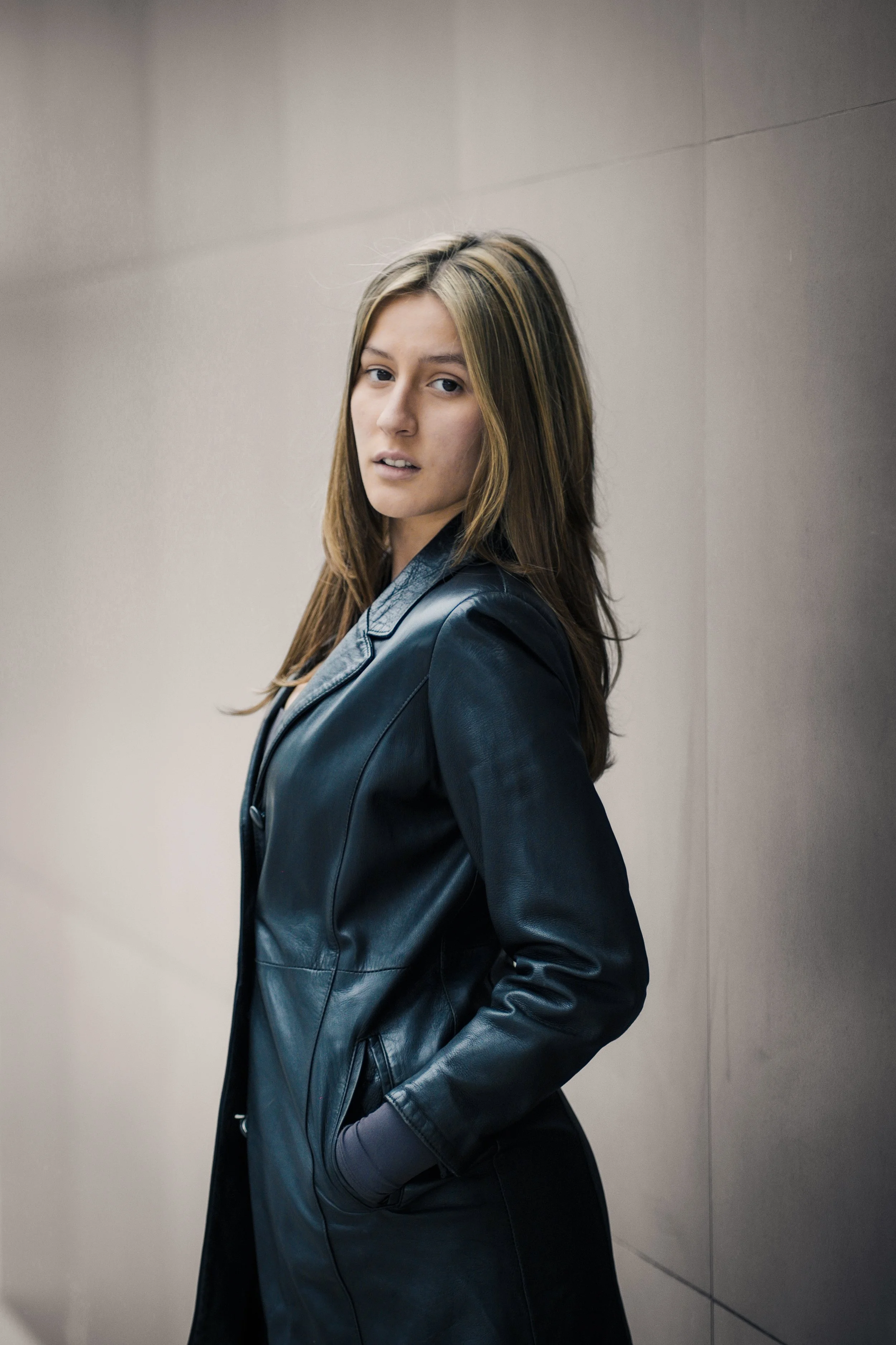 Ragazza con capelli castani, vestita con una giacca in pelle, che posa contro un muro beige.