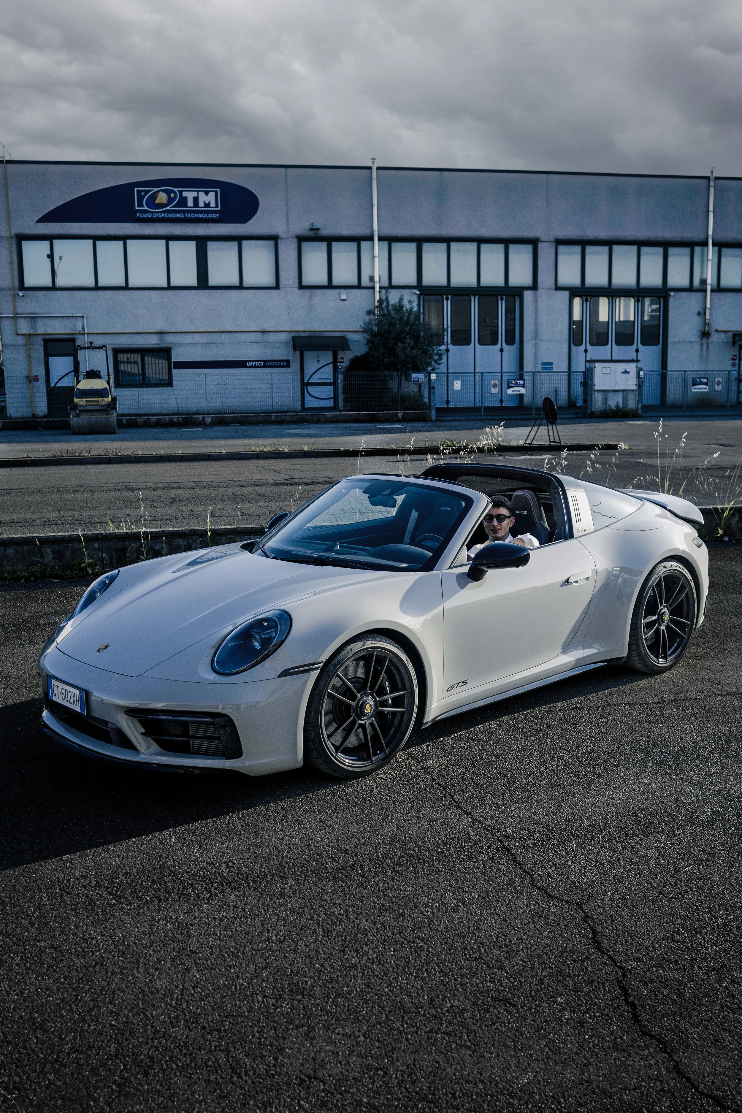 Una vettura decappottabile bianca Porsche Boxster con due persone a bordo, parcheggiata su una strada con un edificio industriale sullo sfondo.