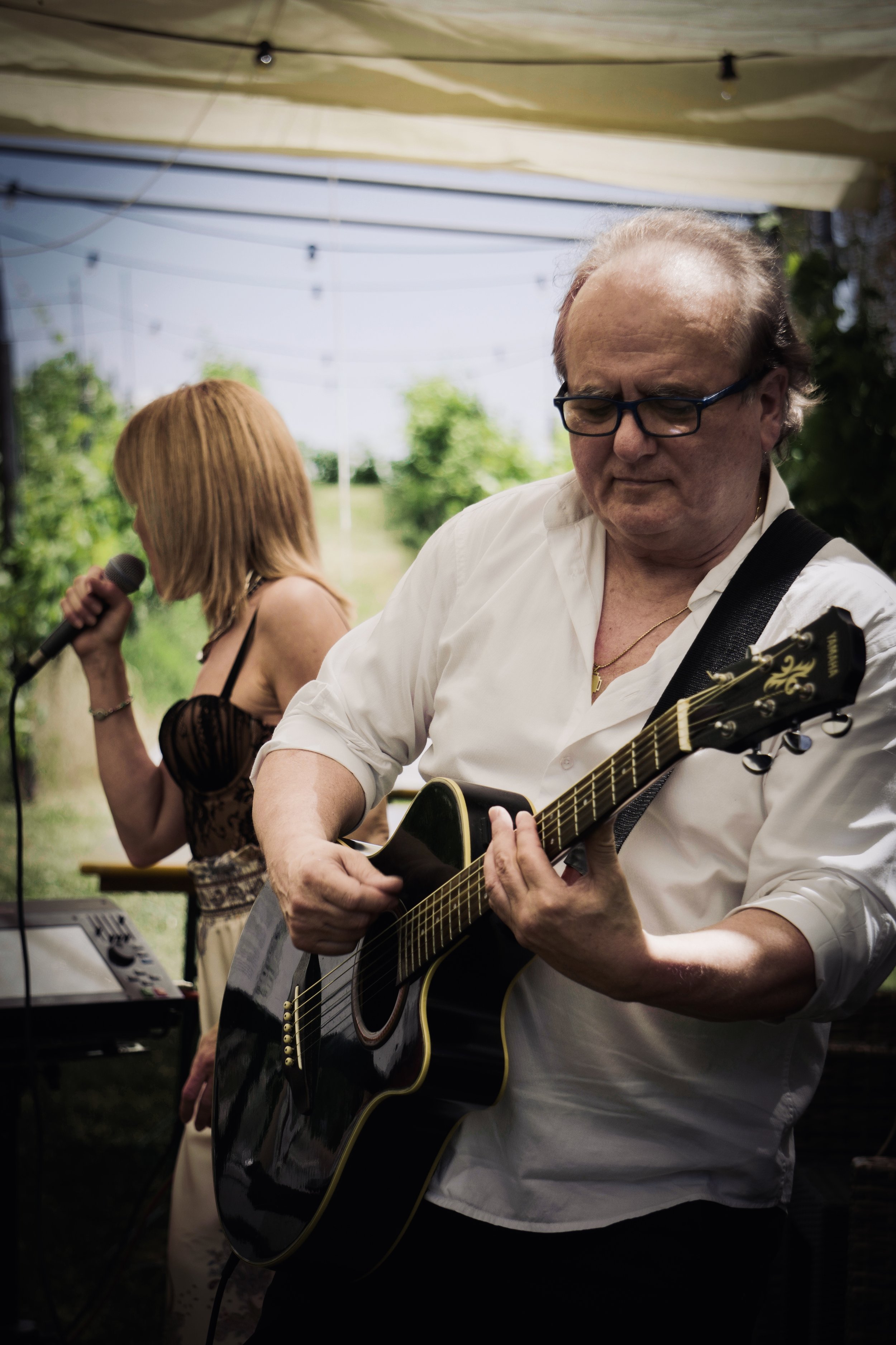 Un uomo che suona chitarra in un evento all'aperto, con una donna che tiene un microfono sullo sfondo.