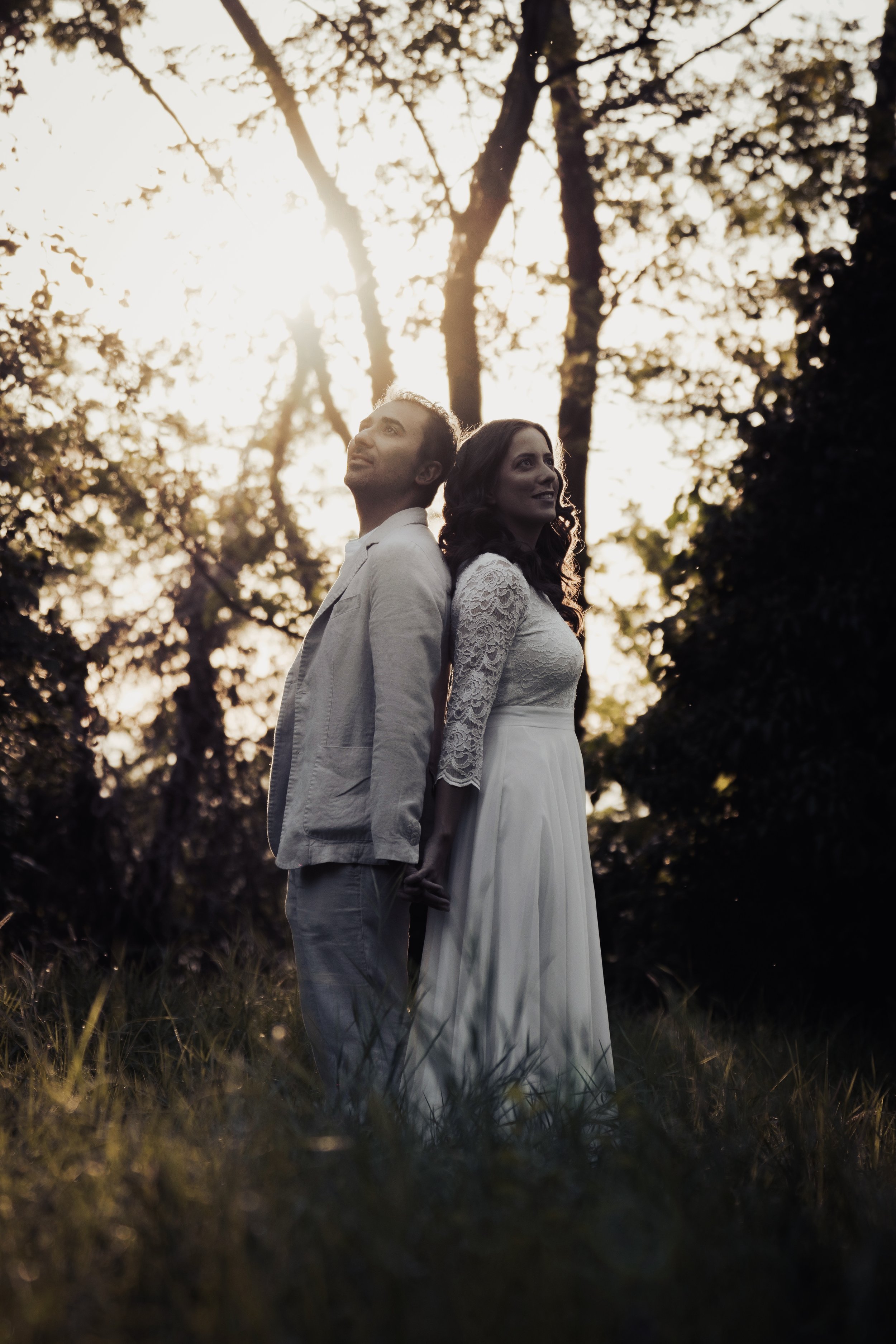 Una coppia di persone in abiti eleganti si tiene per mano in un'atmosfera romantica tra gli alberi, con il sole al tramonto sullo sfondo.