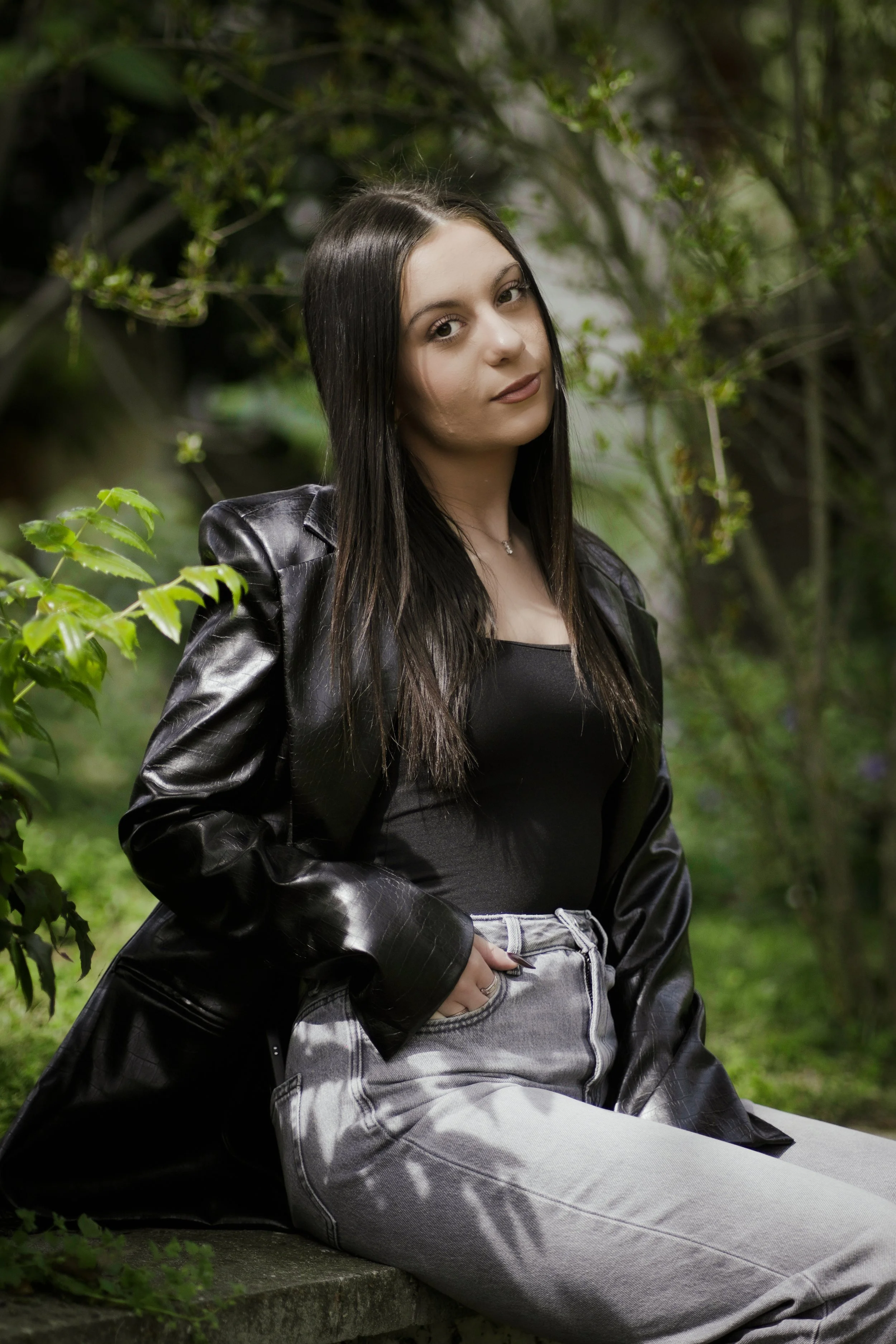 Una ragazza con capelli neri, vestita con una giacca di pelle nera e pantaloni color grigio, seduta su un muretto in un ambiente naturale con foglie verdi.