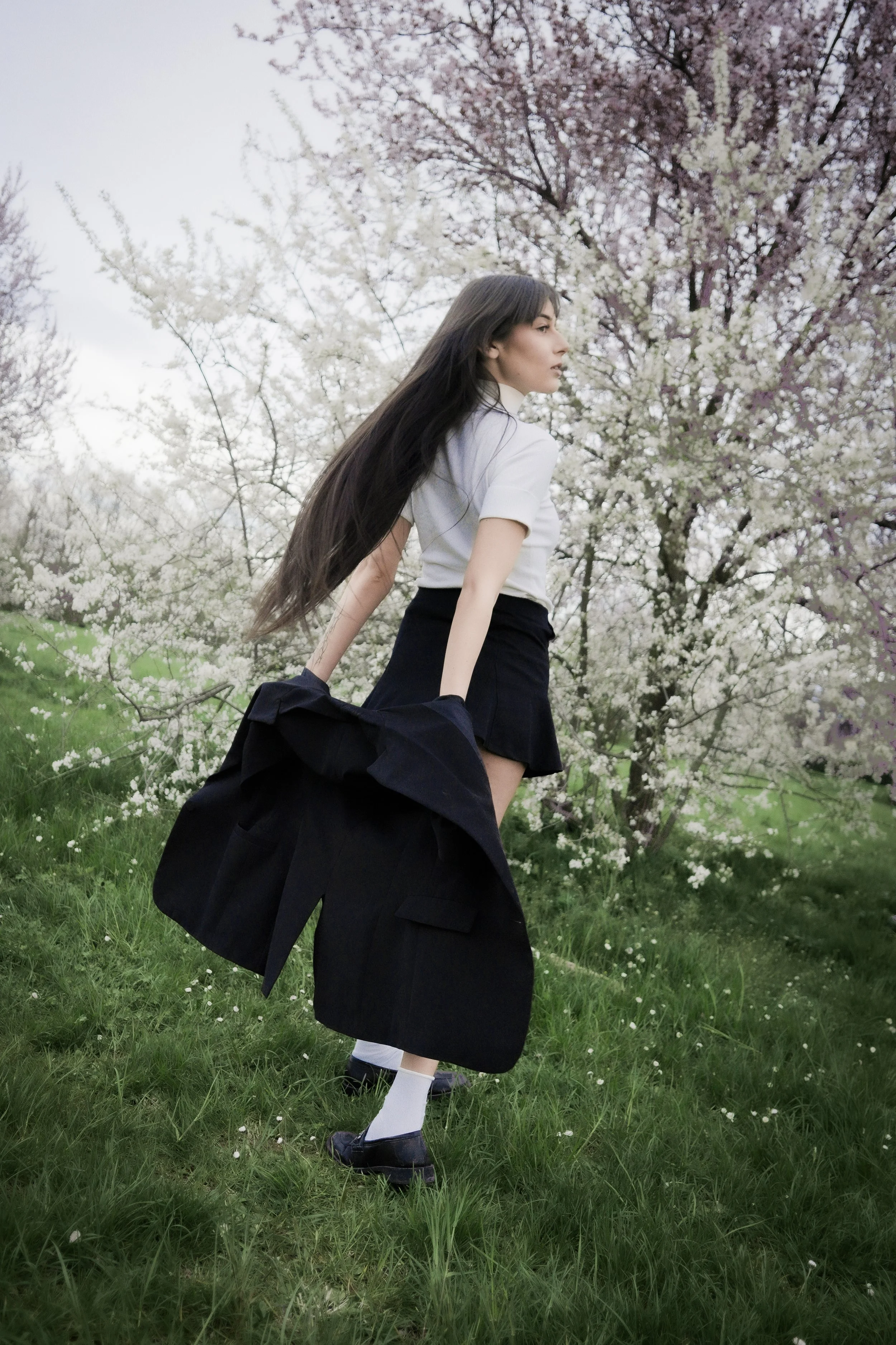 Una donna con capelli lunghi e neri, che indossa una maglietta bianca, una gonna nera e scarpe nere con calze bianche, si trova in un prato con alberi in fiore rosa e bianco.