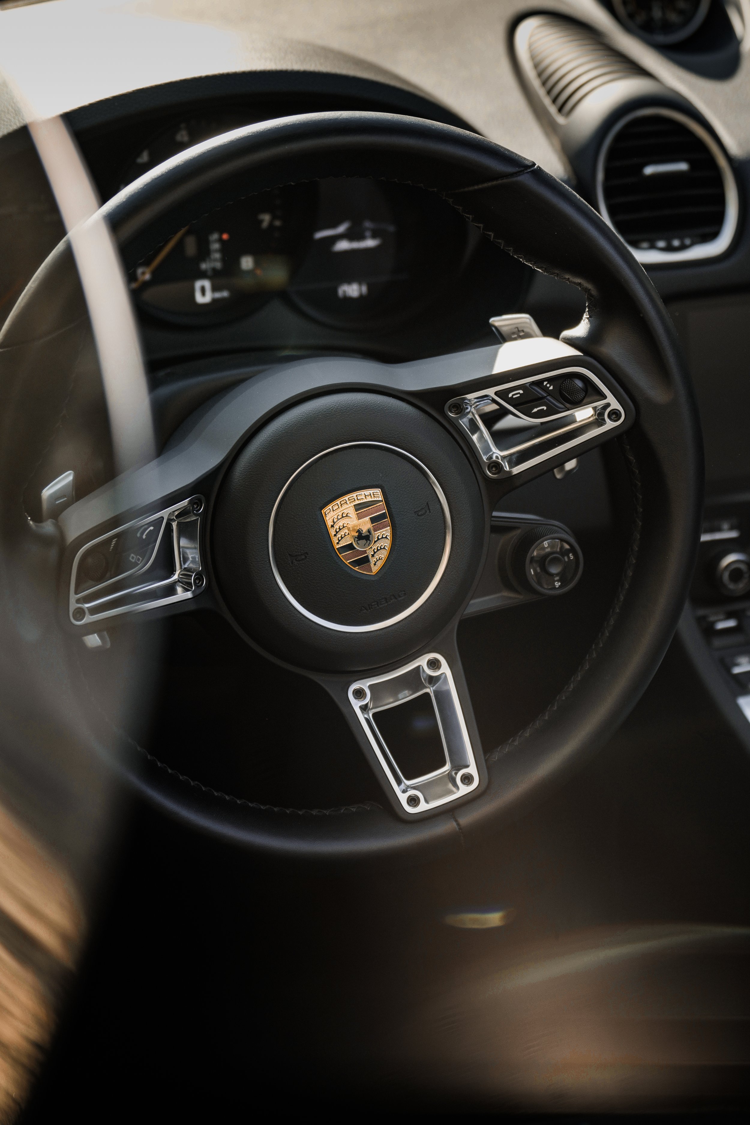 Volante di un'auto Porsche con logo al centro, volante in pelle nera, e alcuni controlli ai lati.