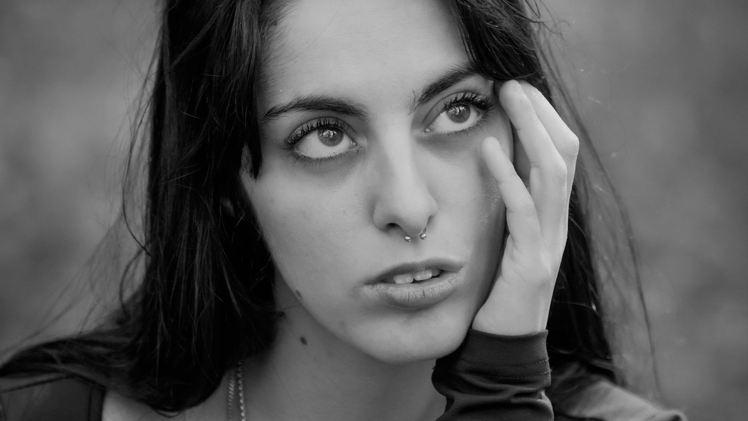 Ragazza con capelli scuri, labbra leggermente socchiuse, con piercing al naso, che tocca il lato del volto con una mano, in bianco e nero.