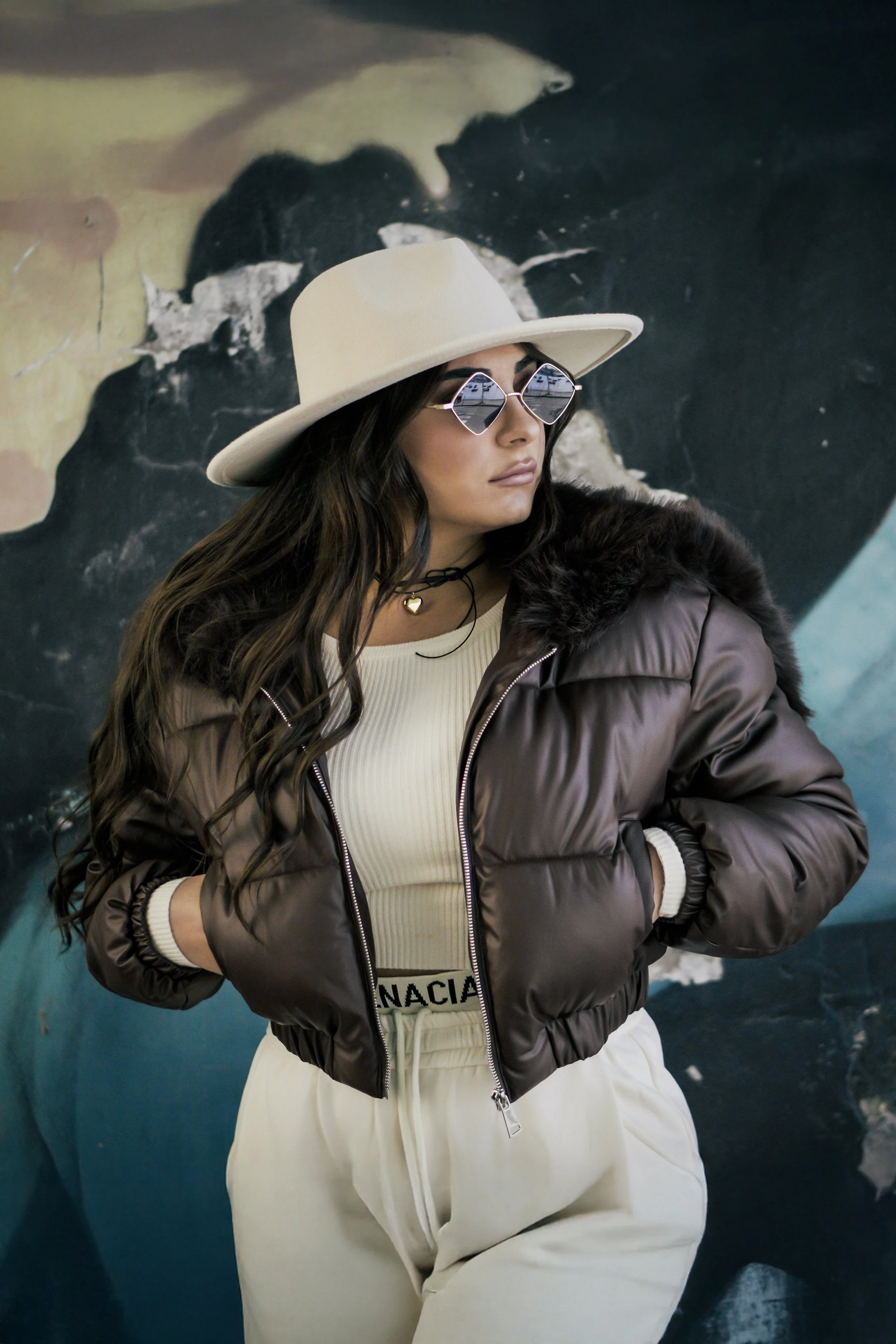 Donna con cappello bianco, occhiali a specchio e giacca imbottita marrone, posa con le mani in tasca davanti a parete di graffiti