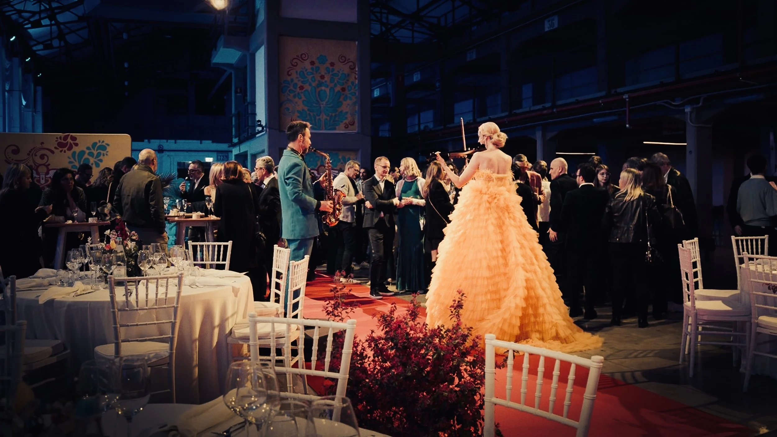 Sfilata di moda o evento artistico con modello che indossa un abito lungo giallo con fan. Musica dal vivo con una band che suona strumenti jazz, pubblico elegante che si intrattiene in un ambiente interno con decorazioni colorate.