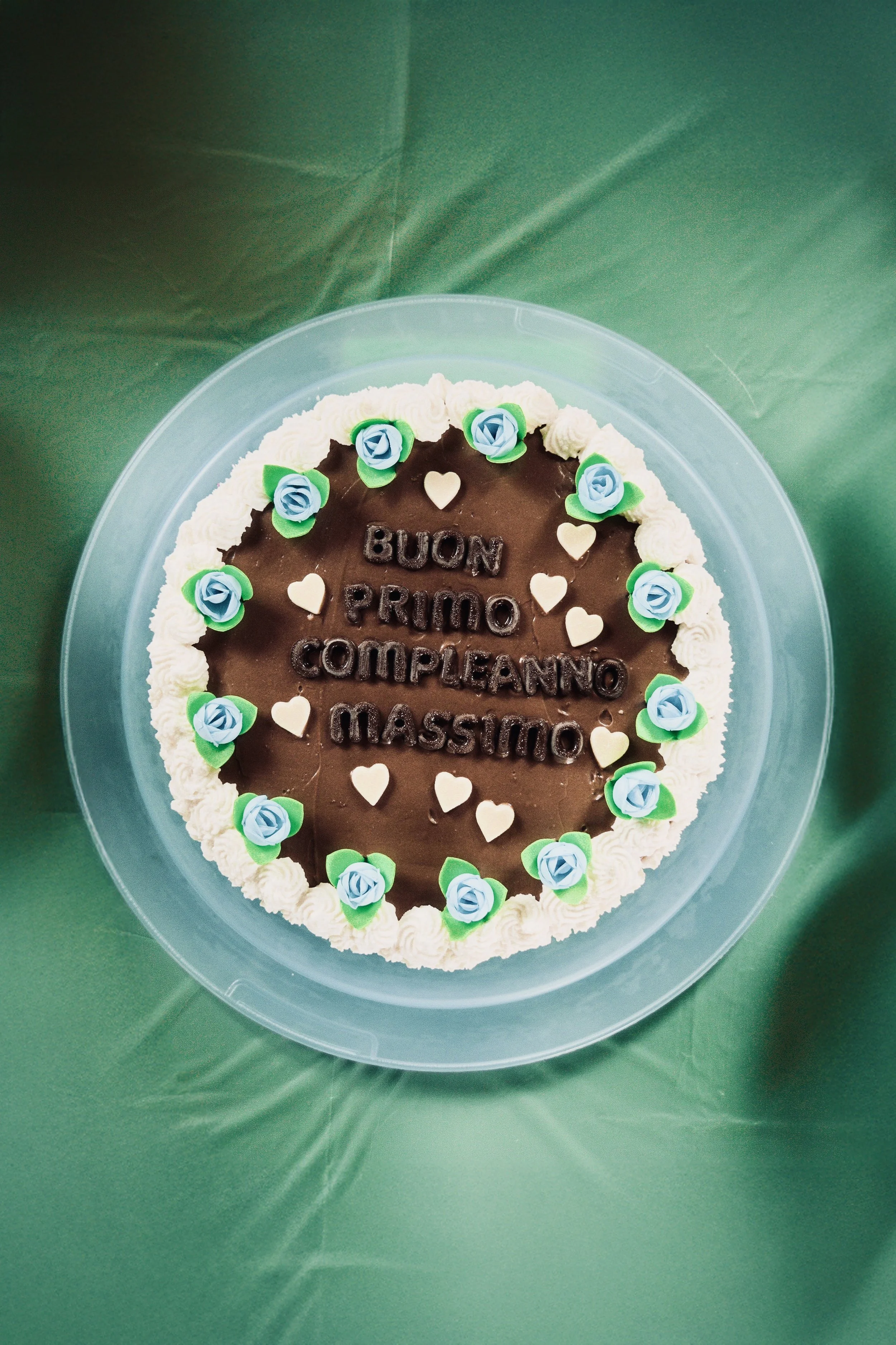 Torta di cioccolato con decorazioni di piccole rose blu e foglie verdi, scritto in cioccolato con la frase in italiano 'Buon primo compleanno massimo', circondata da piccoli cuori bianchi.