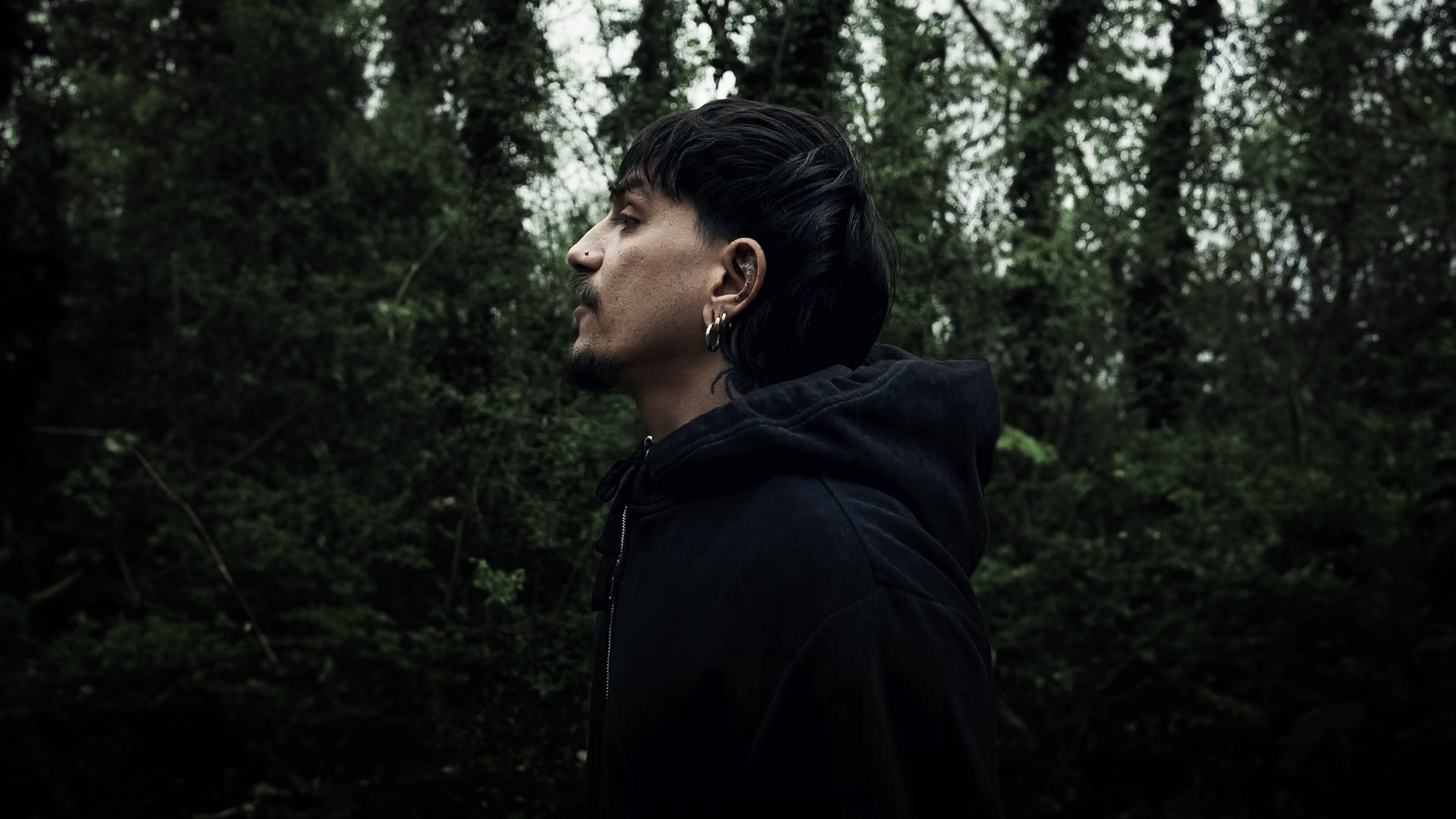 Ragazzo con capelli neri lunghi e barba, indossa cappello e orecchini, guarda di lato in un bosco.