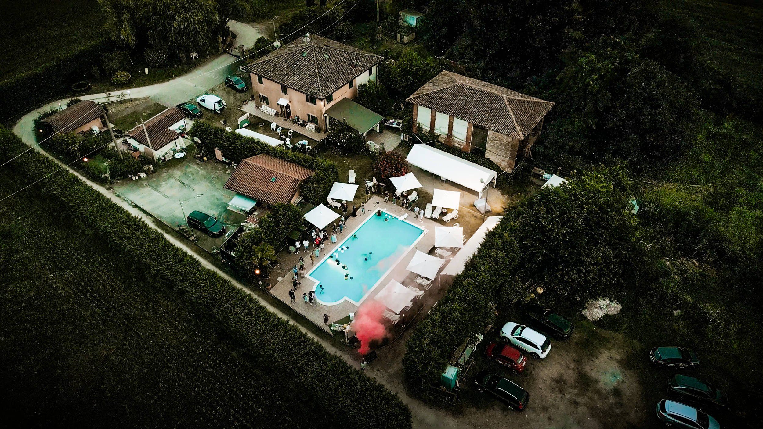 Aerea con piscina e persone che si divertono, circondata da case e auto parcheggiate, con un gruppo di persone che usa del colorato fumo
