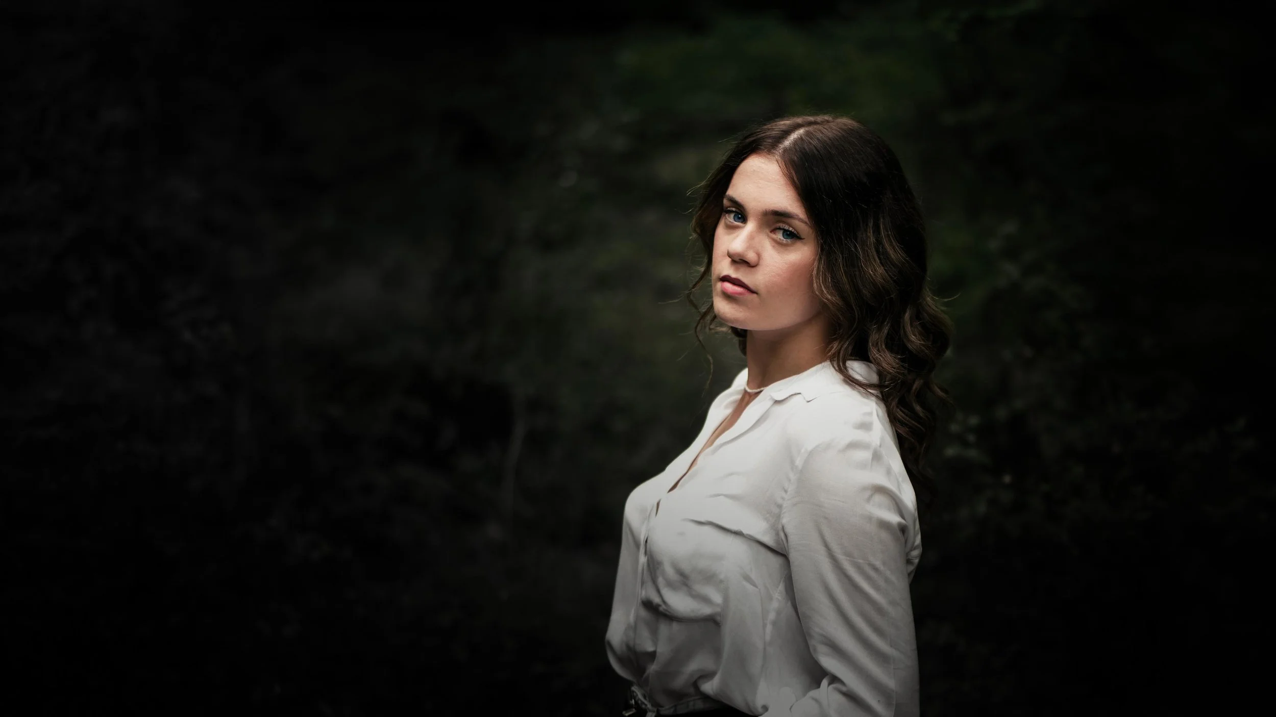Ragazza con capelli castani e occhi azzurri indossa una camicia bianca, sfondo scuro e naturale.