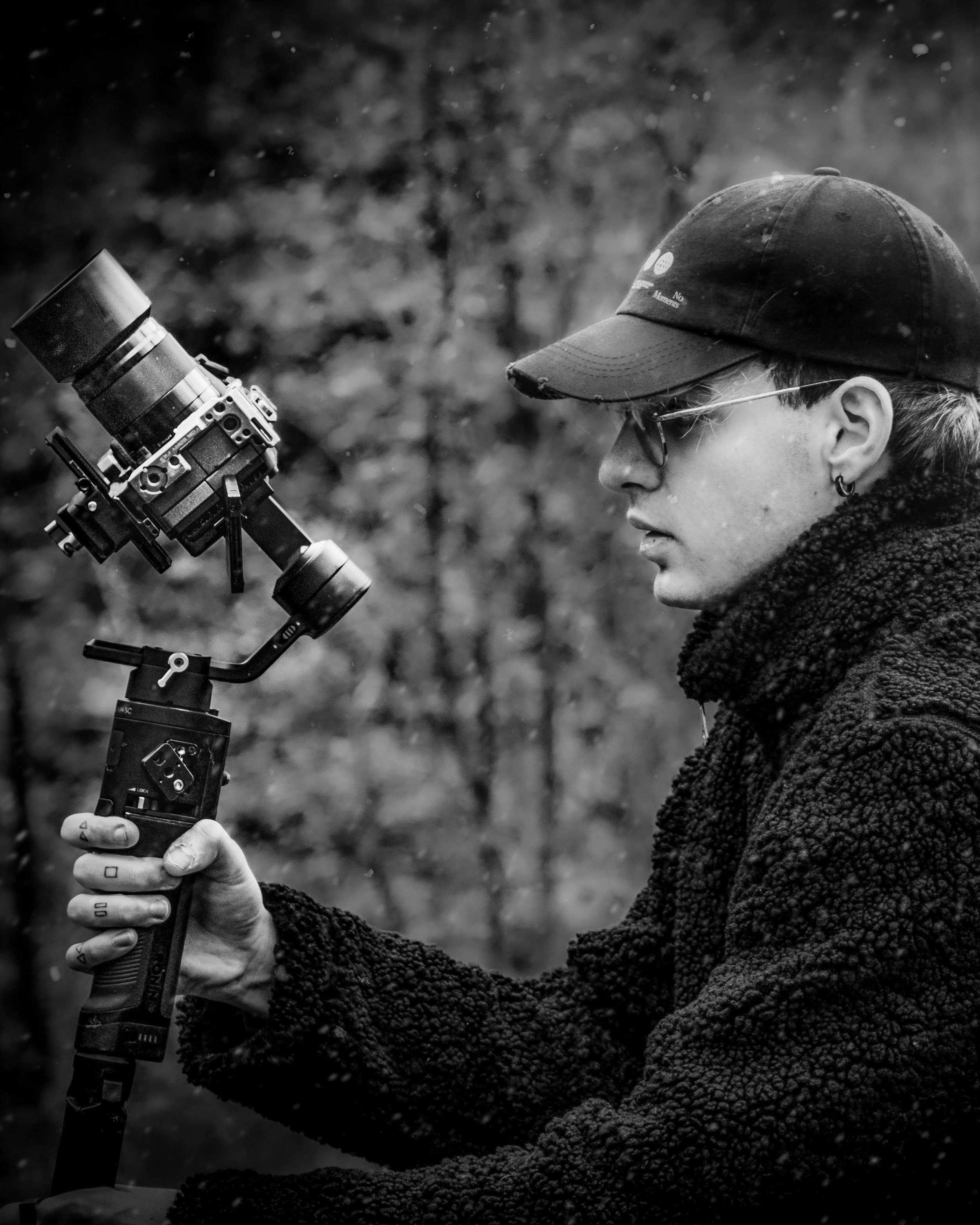 Una persona con cappellino, occhiali e orecchini, che tiene stabilizzato un teleobiettivo con un gimbal in un ambiente naturale.