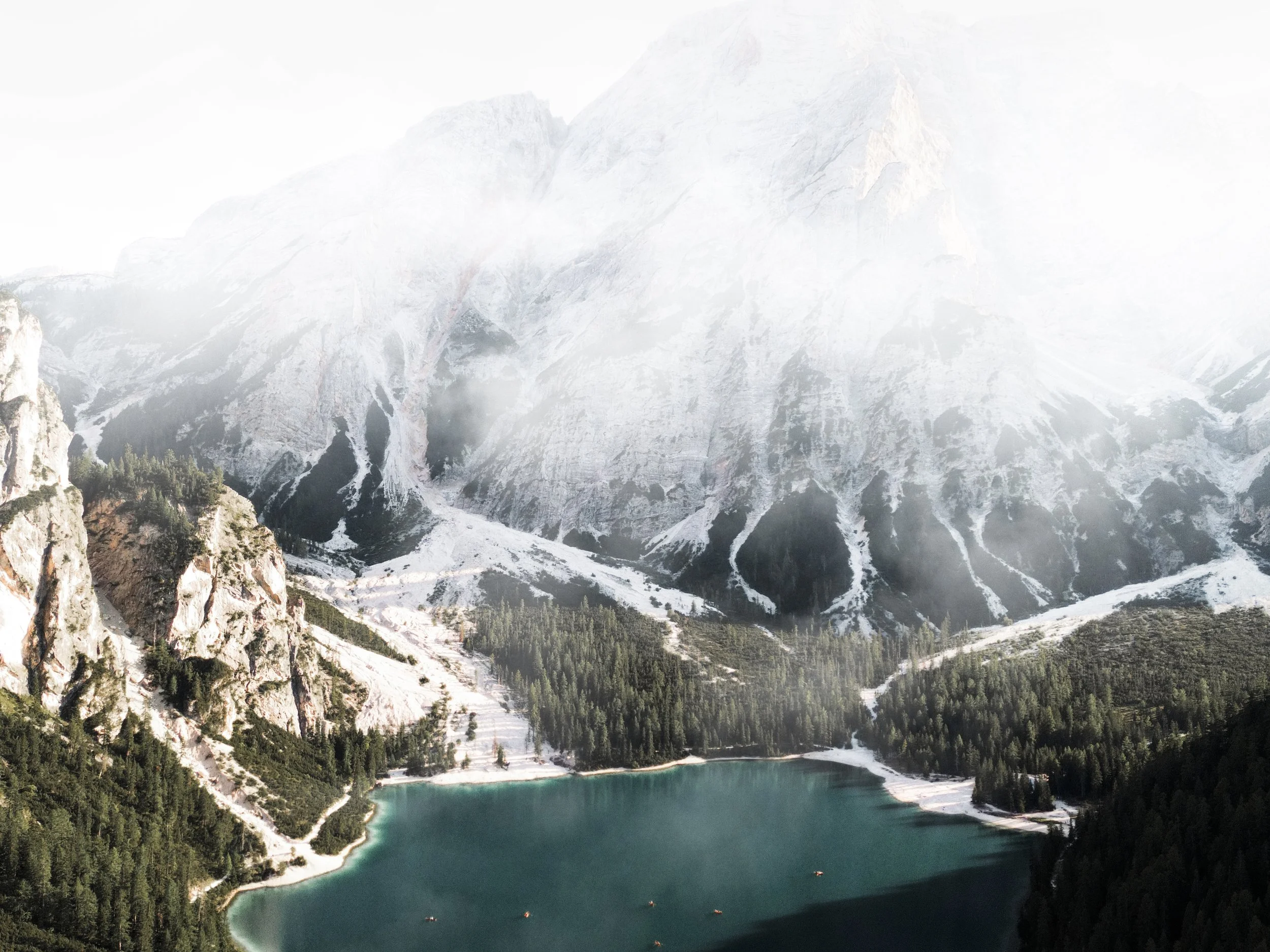 Paesaggio montano con un lago turchese circondato da foreste e montagne innevate in lontananza.