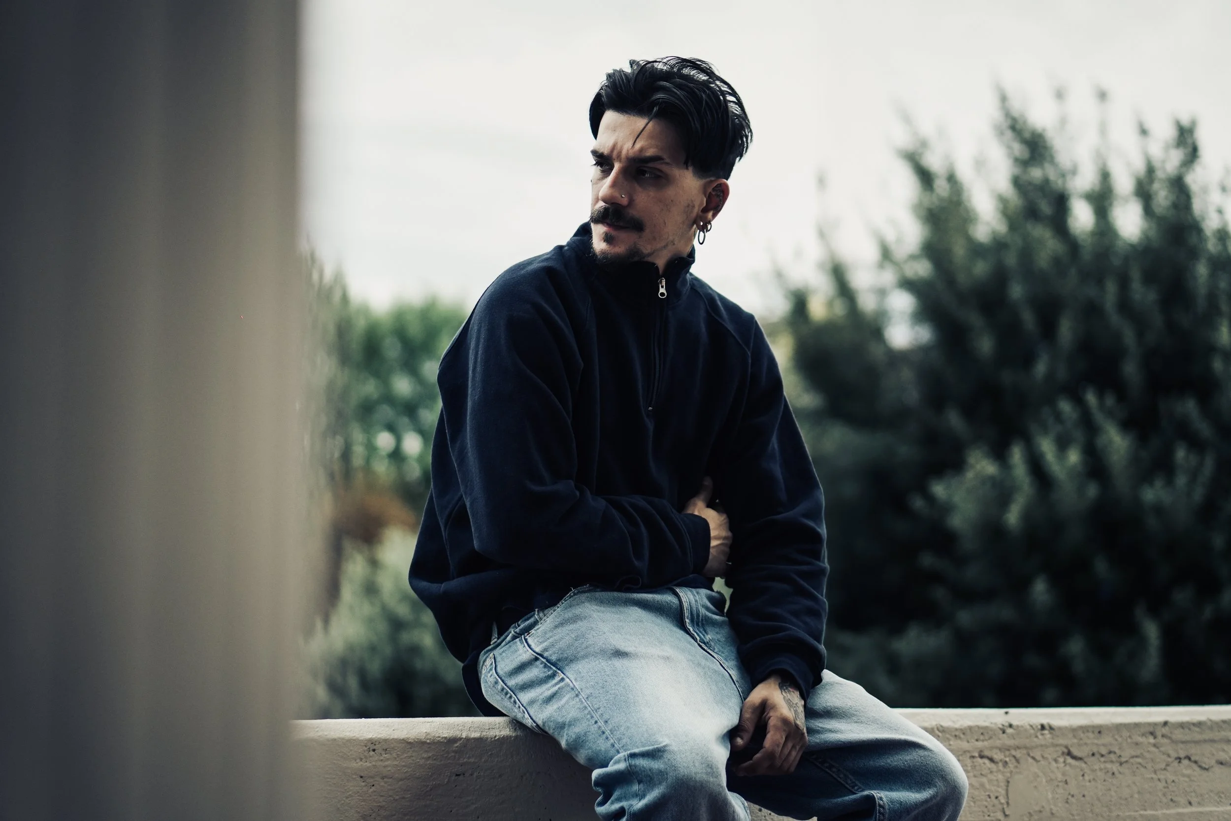 Un uomo con capelli neri, giacca nera e jeans, seduto su un muretto, guarda a sinistra, con uno sfondo di alberi e cielo.