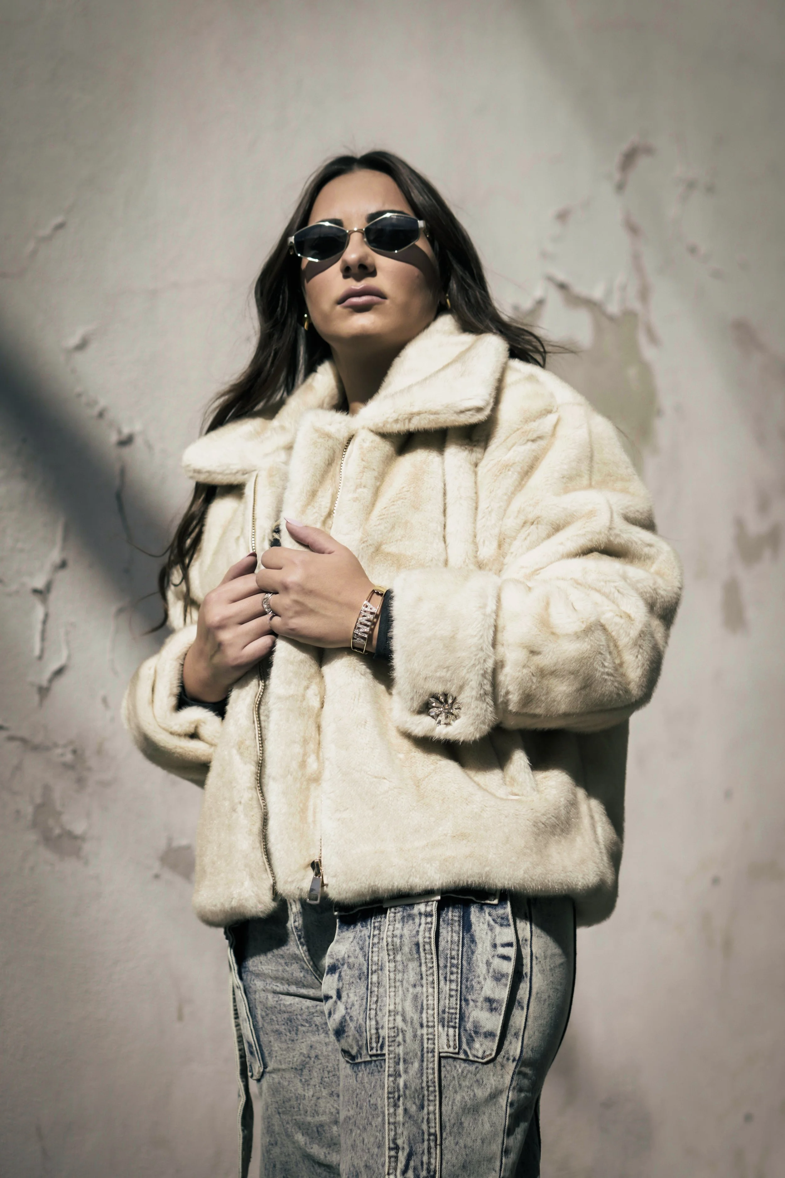 Donna con occhiali da sole scuri, cappotto di pelliccia color crema, e jeans, posando contro un muro.