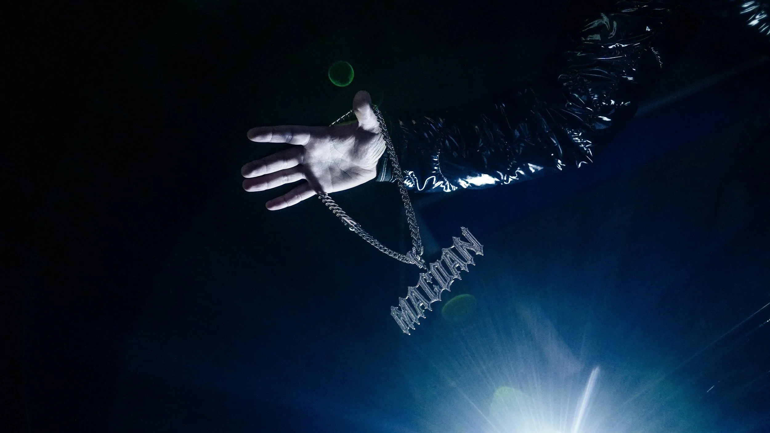 Mano con pelle scamosciata che tiene una collana con la parola "NUTTHIN" pendente, sfondo scuro con alcuni riflessi.