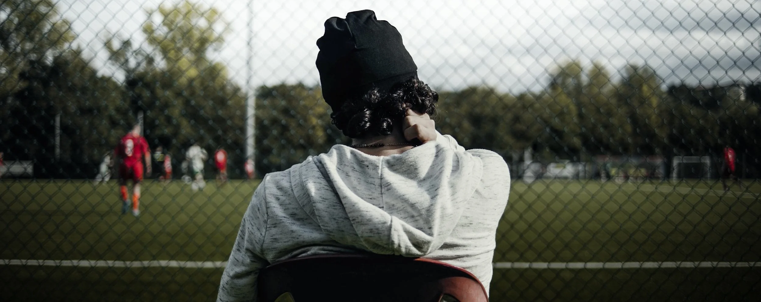 Una persona con cappello nero osserva una partita di calcio attraverso una rete al campo.