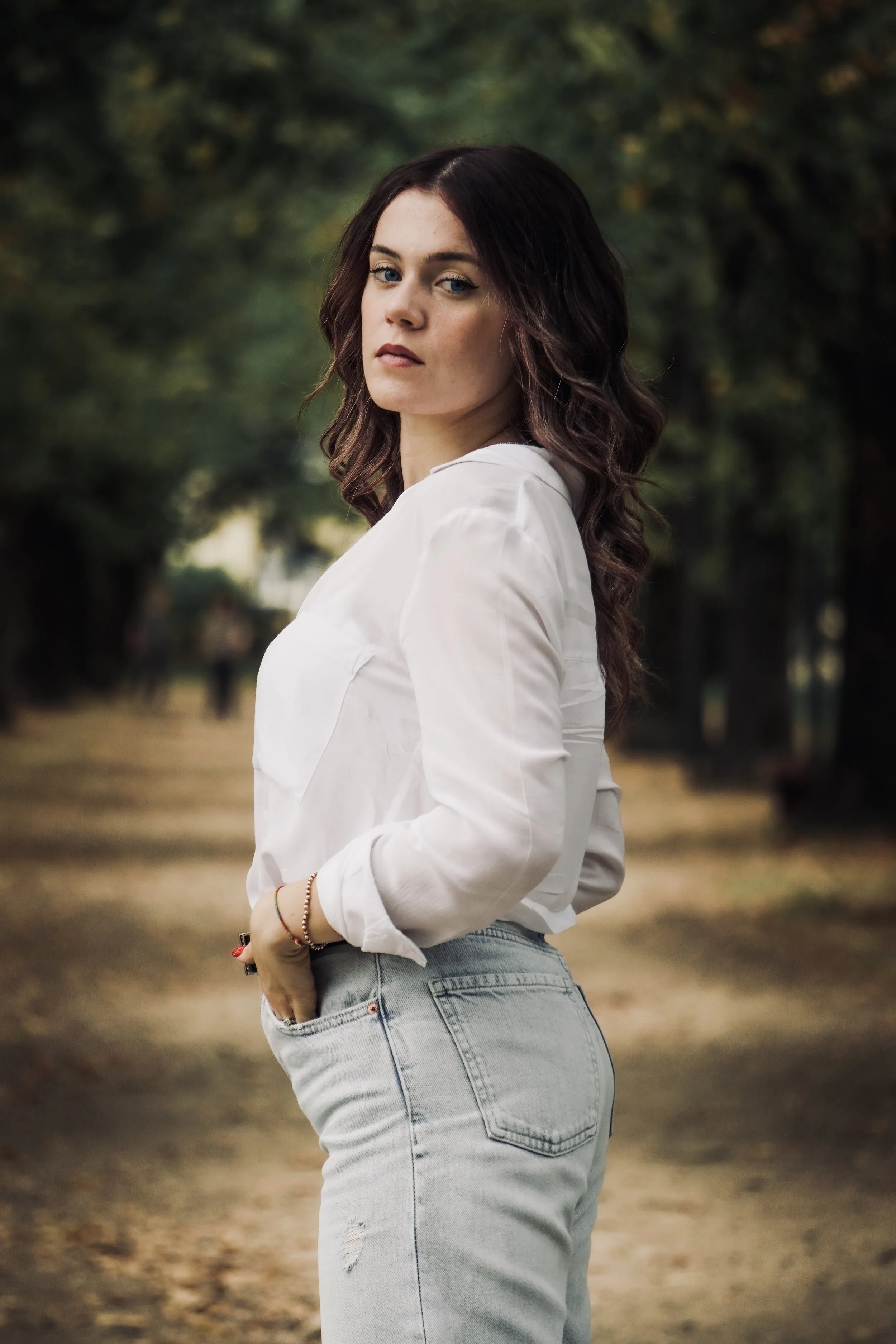 Una donna dai capelli ricci e vestita con camicia bianca e jeans chiari si trova in un parco circondato dagli alberi.