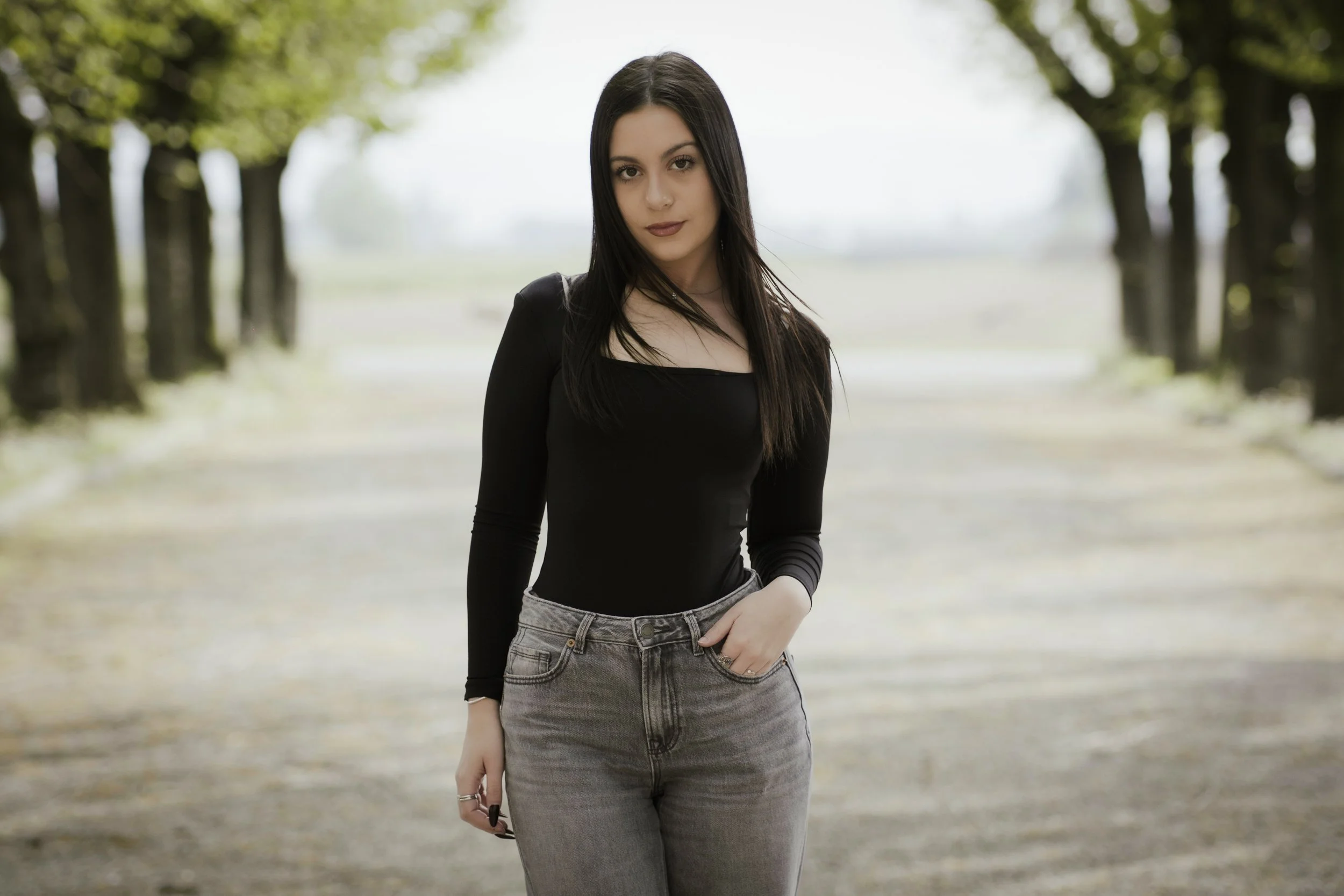 Ragazza con capelli neri, maglietta nera a maniche lunghe e pantaloni grigi, che si trova su una strada circondata da alberi.