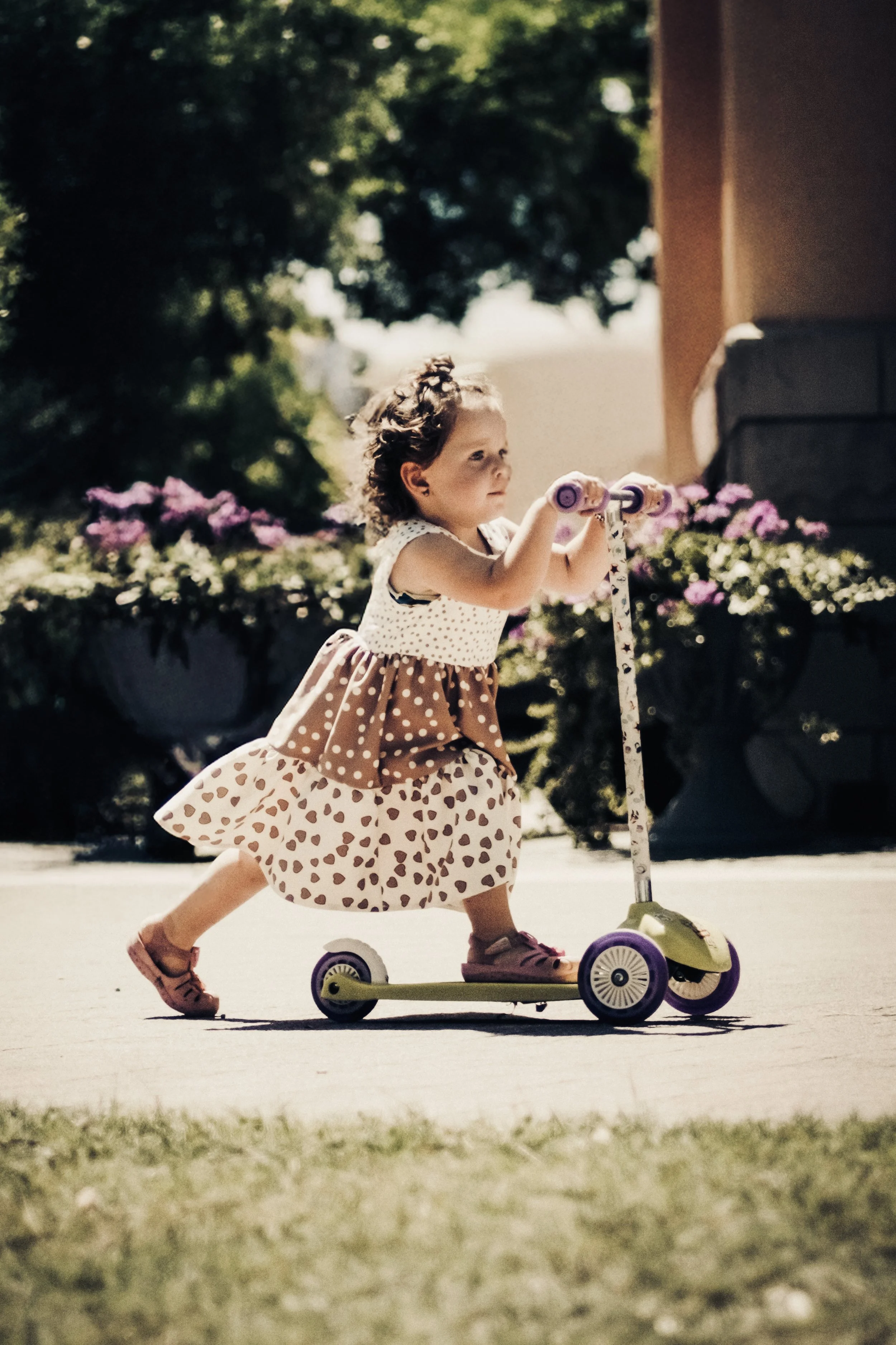 Una bambina in piedi su uno scooter giocattolo rosa e verde, che si muove su una superficie di cemento all'aperto, con fiori viola e piante verdi sullo sfondo.