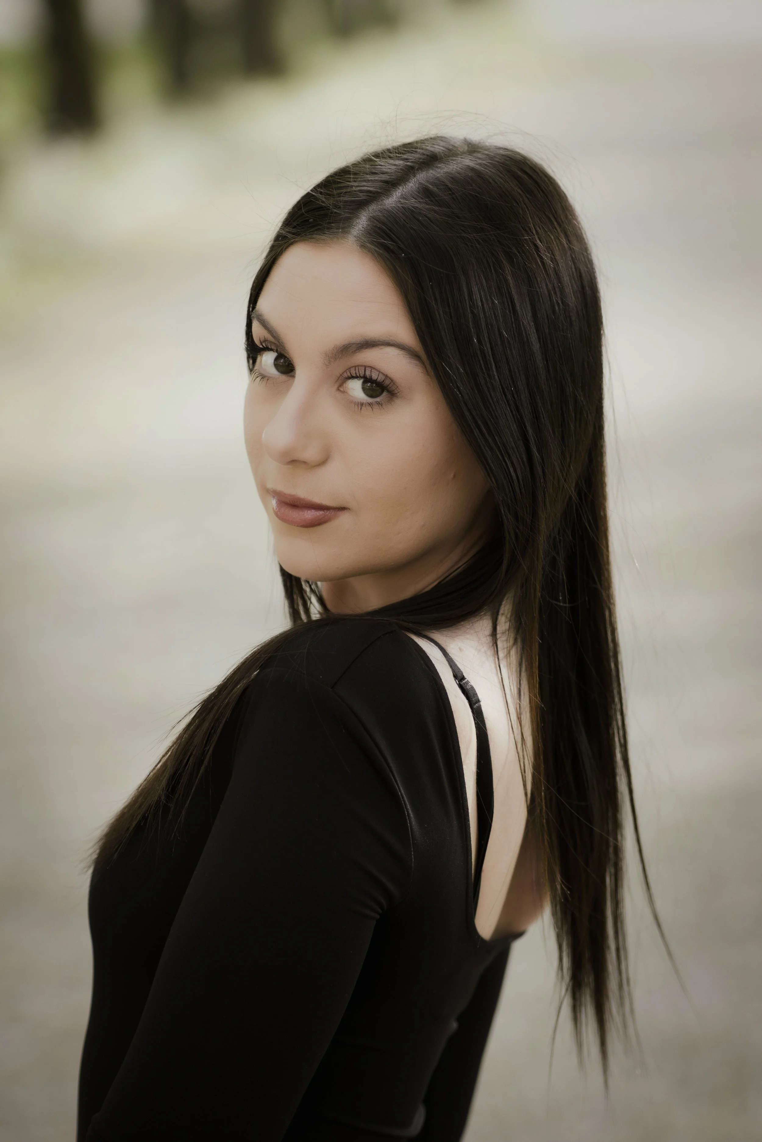 Ragazza con vestito aderente nero, capelli lunghi e castani, sfondo naturale sfocato.