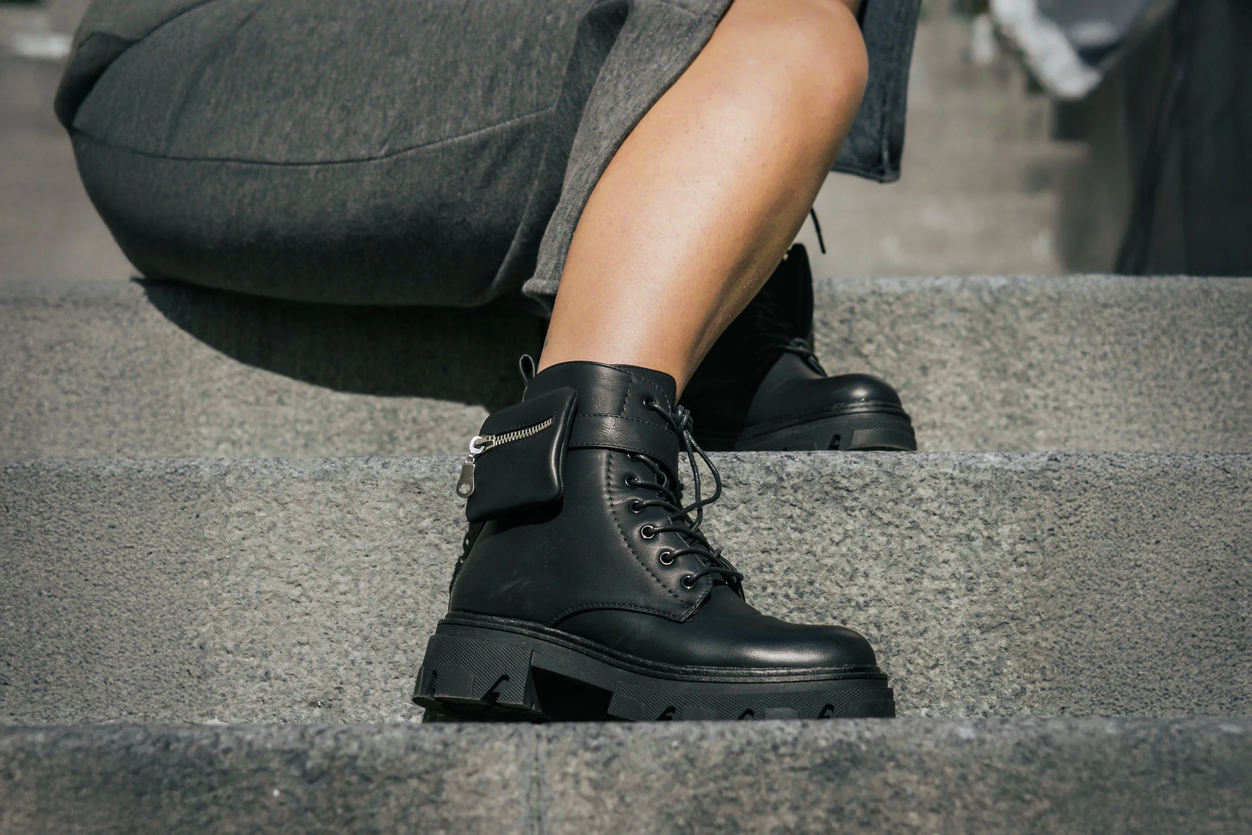 Scarpe nere in pelle con suola spessa, indossate da una persona seduta sulle scale di pietra, con un pantalone grigio e una tasca porta oggetti abbinata alle scarpe.